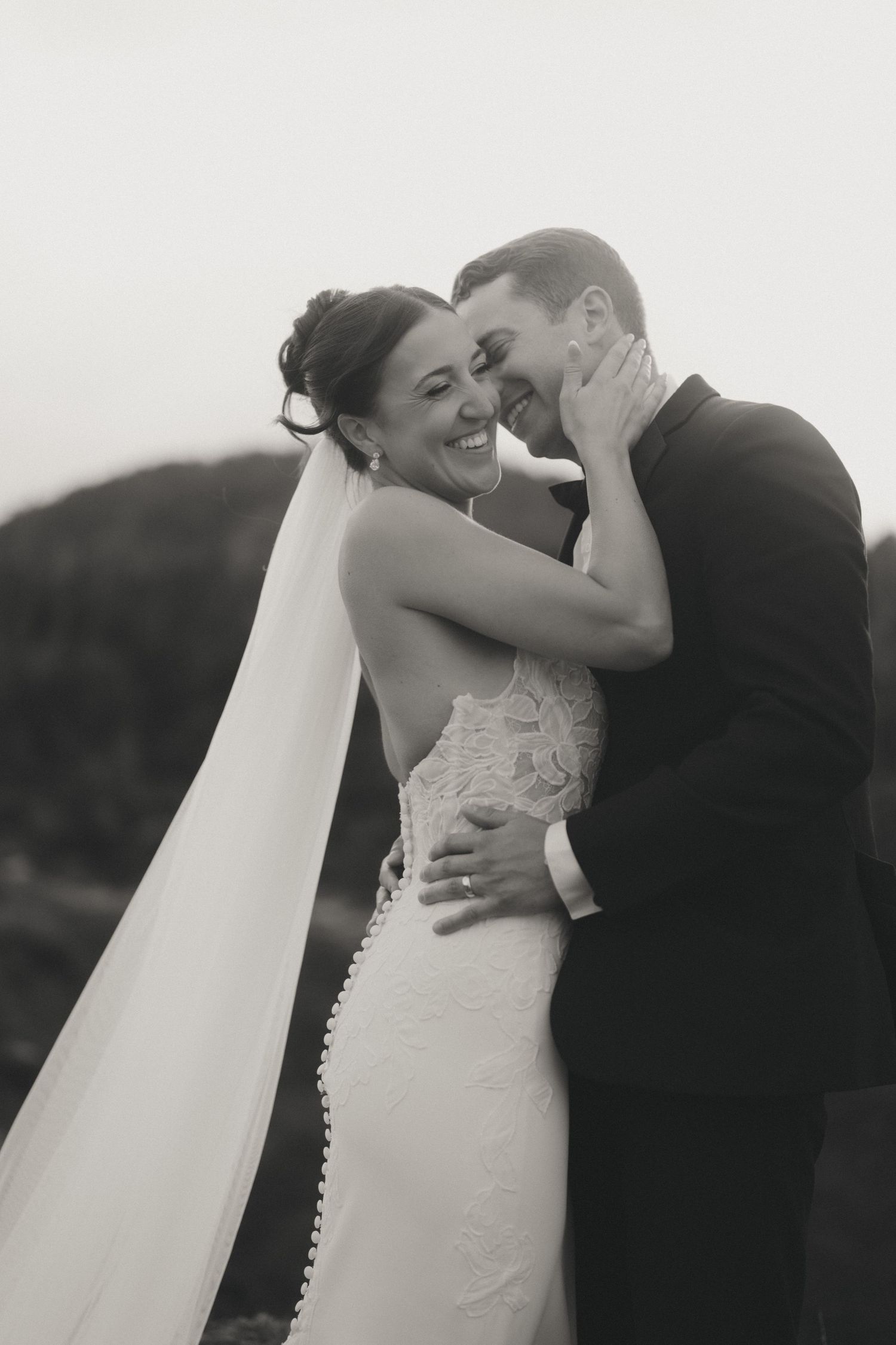 Black and white photo of a smiling bride in a backless lace gown and veil embracing her groom outdoors.