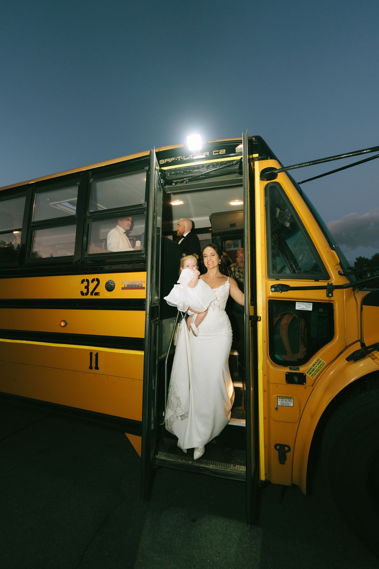 A couple in formal attire poses for wedding photos while entering and exiting a vintage yellow school bus at night.