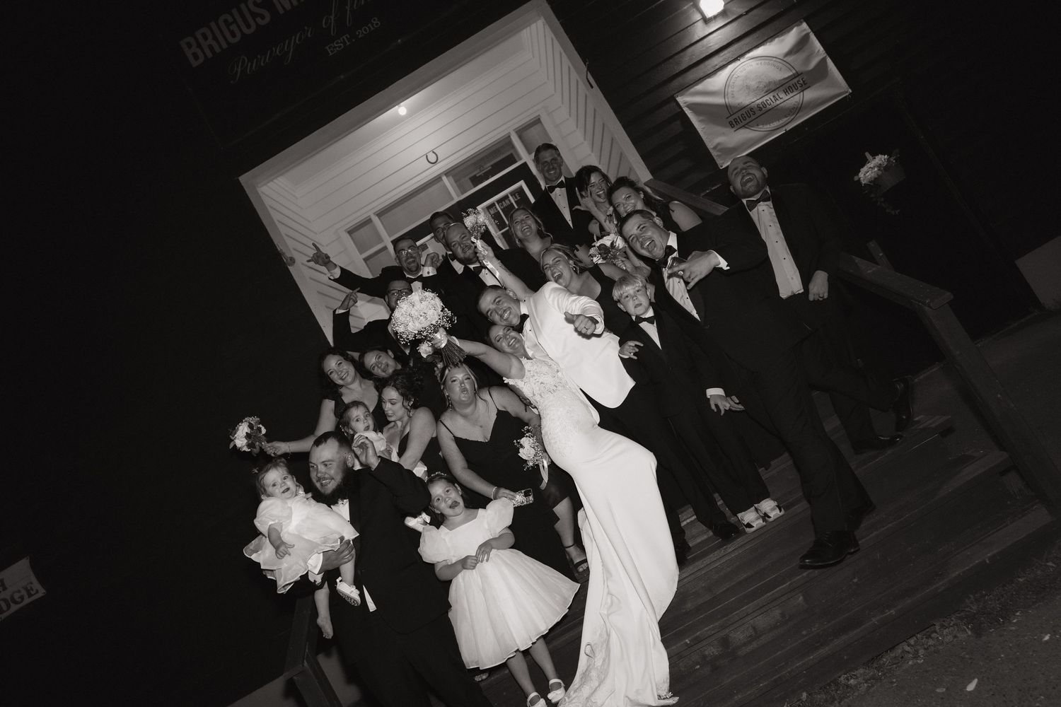 A black and white photo captures guests dancing and celebrating at a wedding reception on a dance floor.