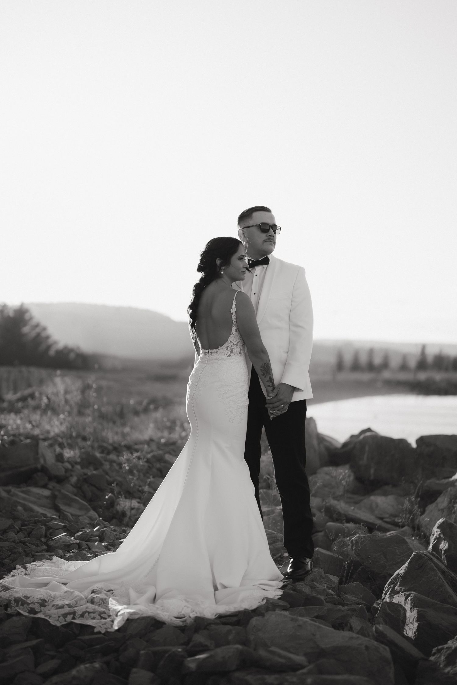 A couple in wedding attire shares an intimate moment on a rocky shore at sunset in black and white.