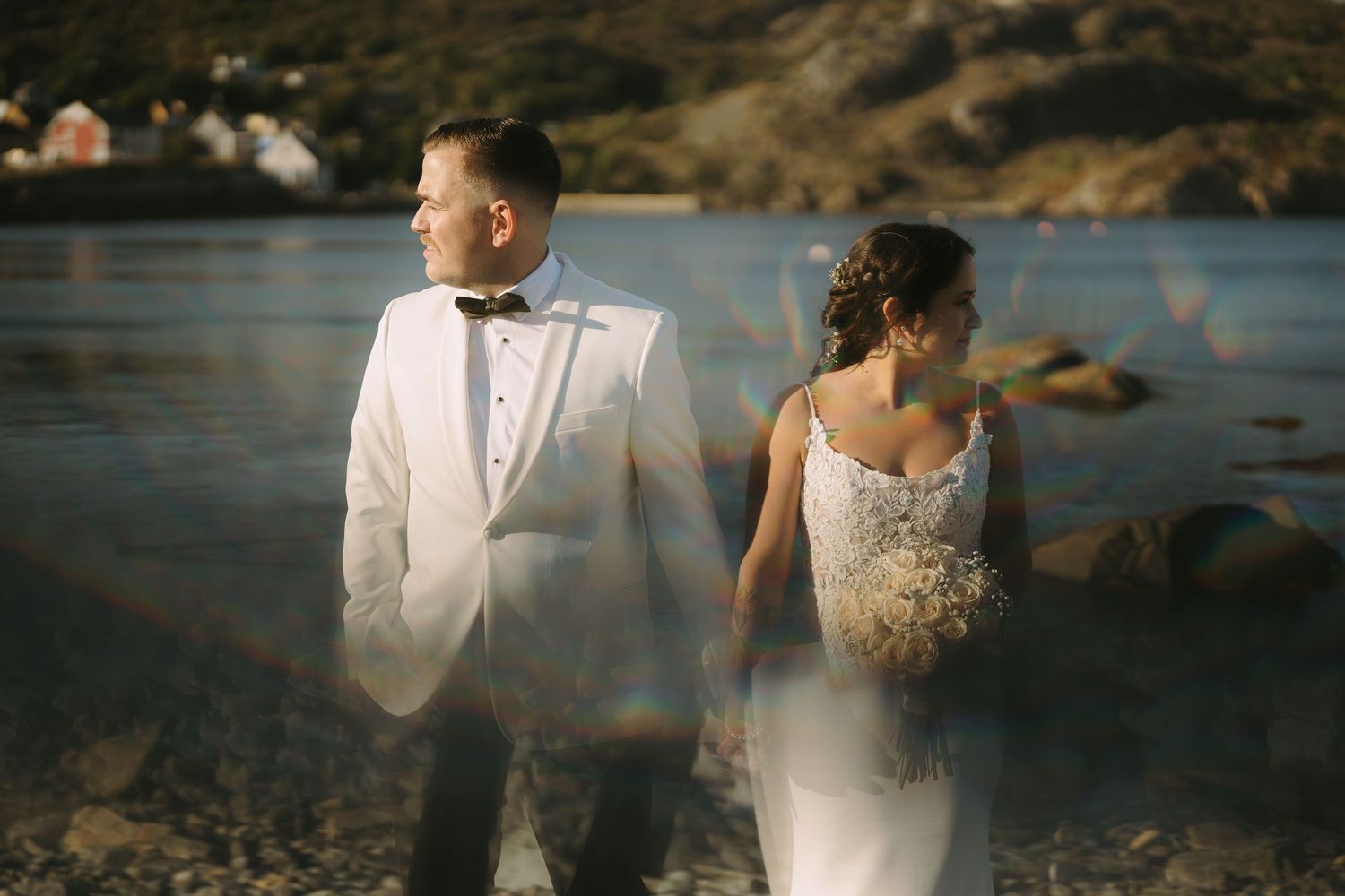 A couple in wedding attire stands by a scenic waterfront at sunset creating a romantic silhouette against the golden light.