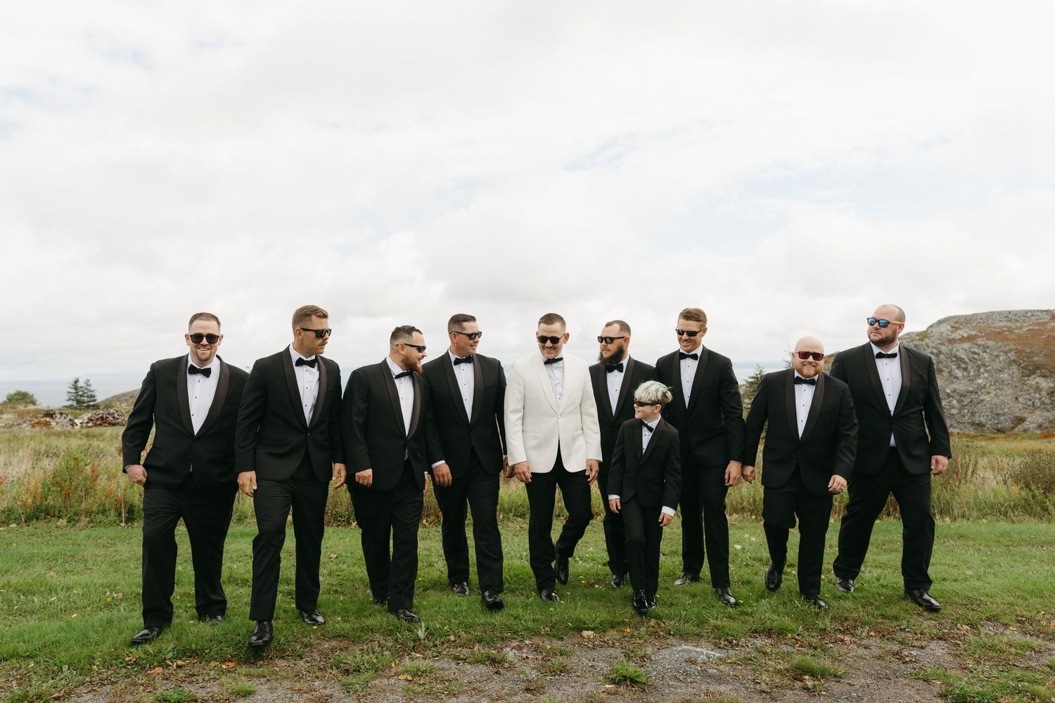 A group of groomsmen in black suits and sunglasses pose together outdoors in a scenic mountain landscape.