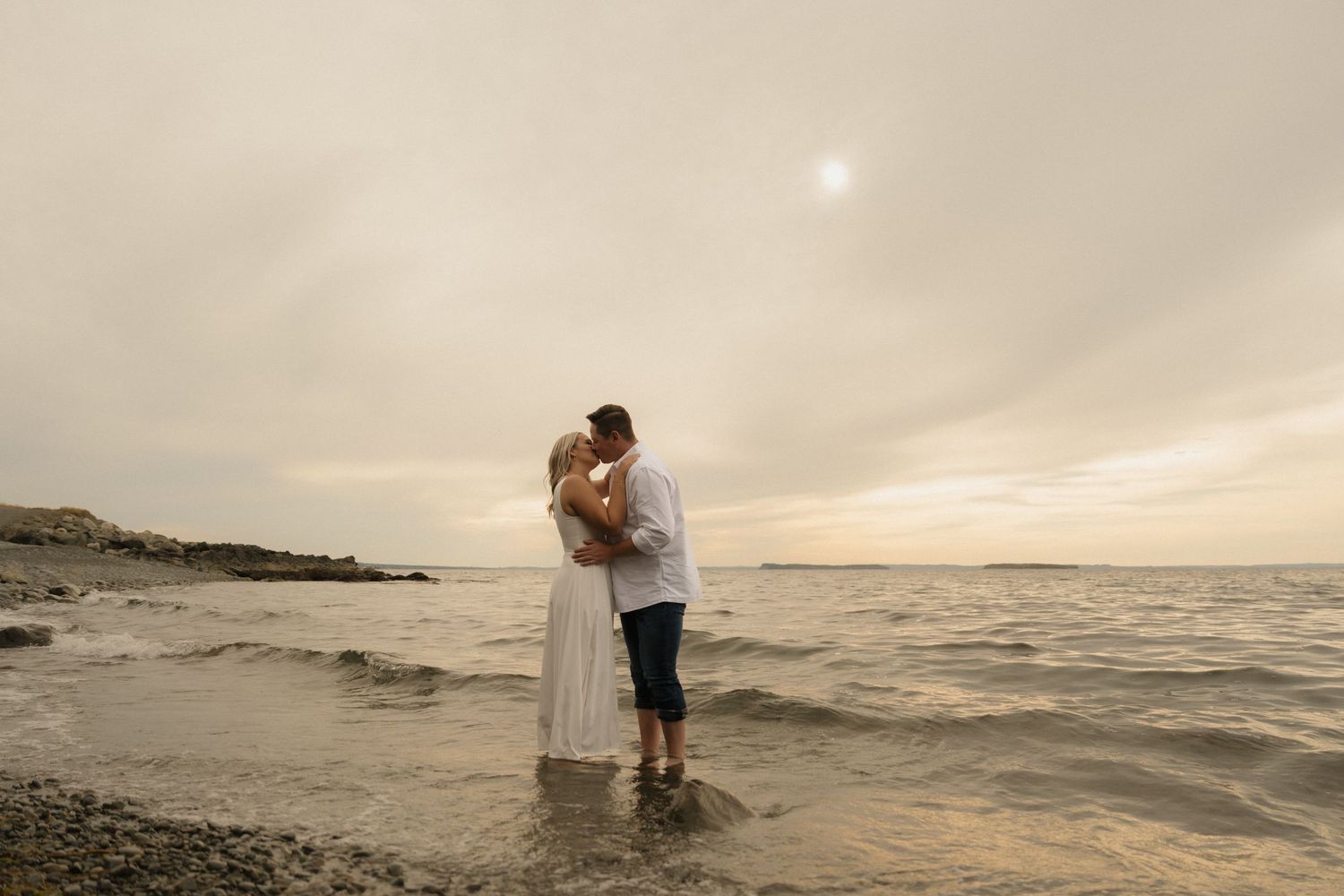 A couple embraces on a serene beach at sunset with gentle waves rolling onto the shore.