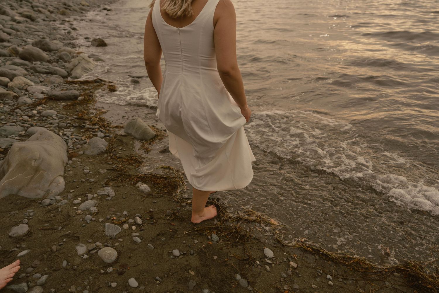 Person in a white dress walks along a rocky shoreline at sunset with rippling water in the background.