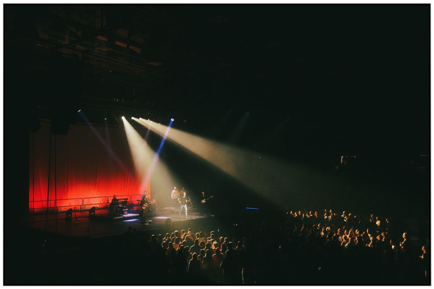 Dramatic spotlights illuminate a dark concert venue with red lighting and a large crowd gathered below.