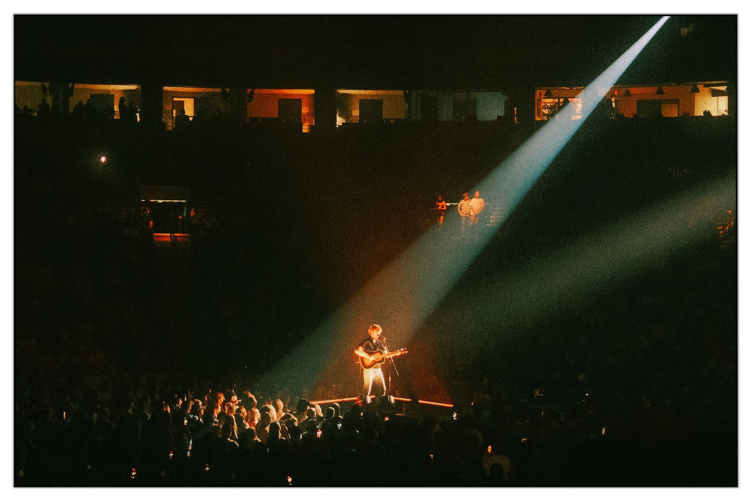 Concert performer stands with arms outstretched on stage under dramatic spotlights while crowd watches from below.