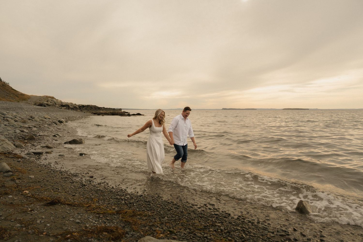A couple walks along a rocky beach at sunset with waves lapping at the shore.