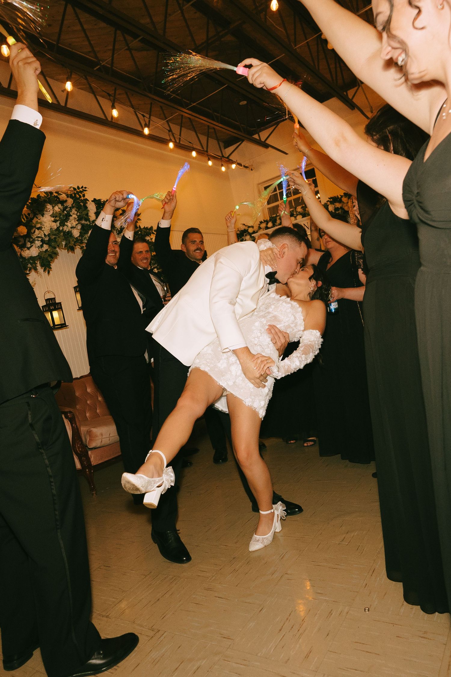 Couple shares romantic dip kiss on dance floor at wedding reception while guests cheer with raised arms in celebration.