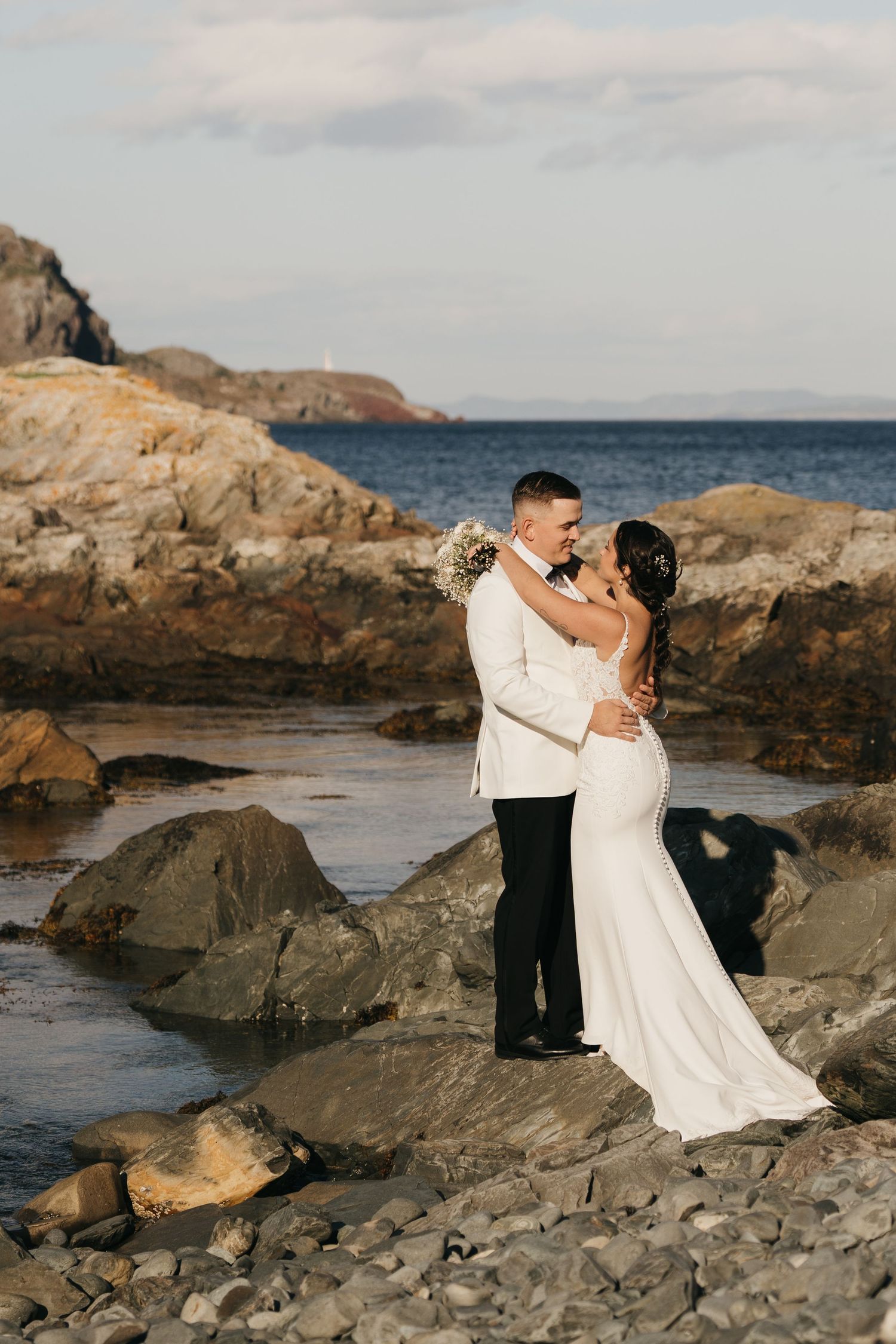 A couple embraces on rocky coastal cliffs during sunset with ocean waves and mountains in the background.