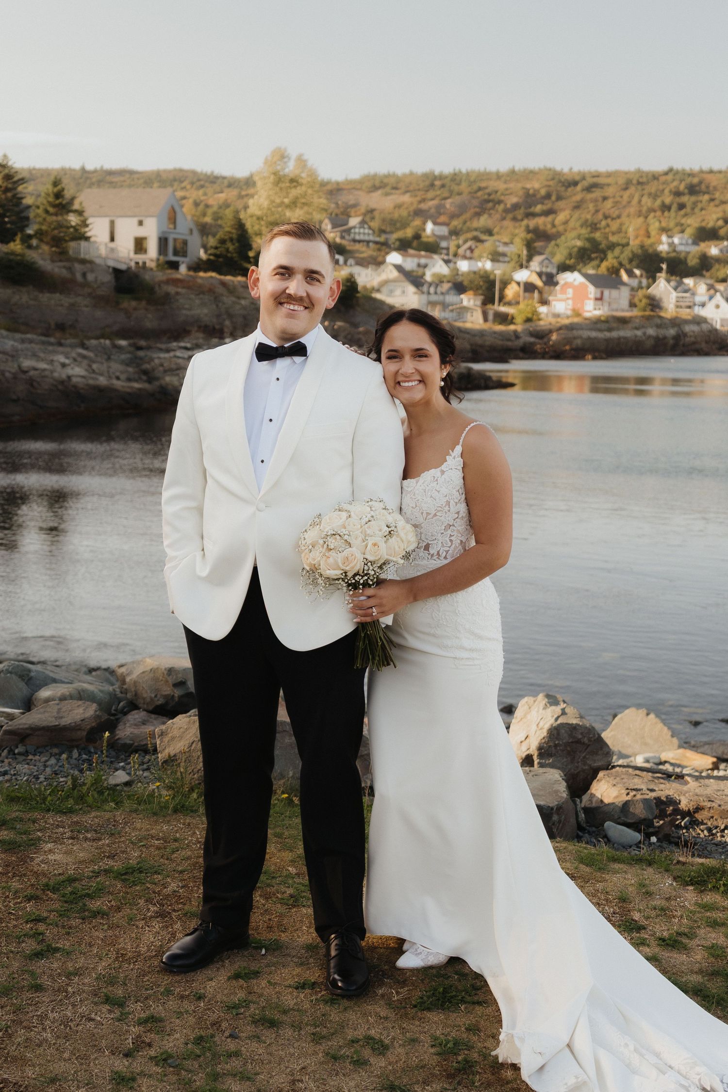 A couple in formal wedding attire poses by a waterfront with rocks and trees in the background at sunset.