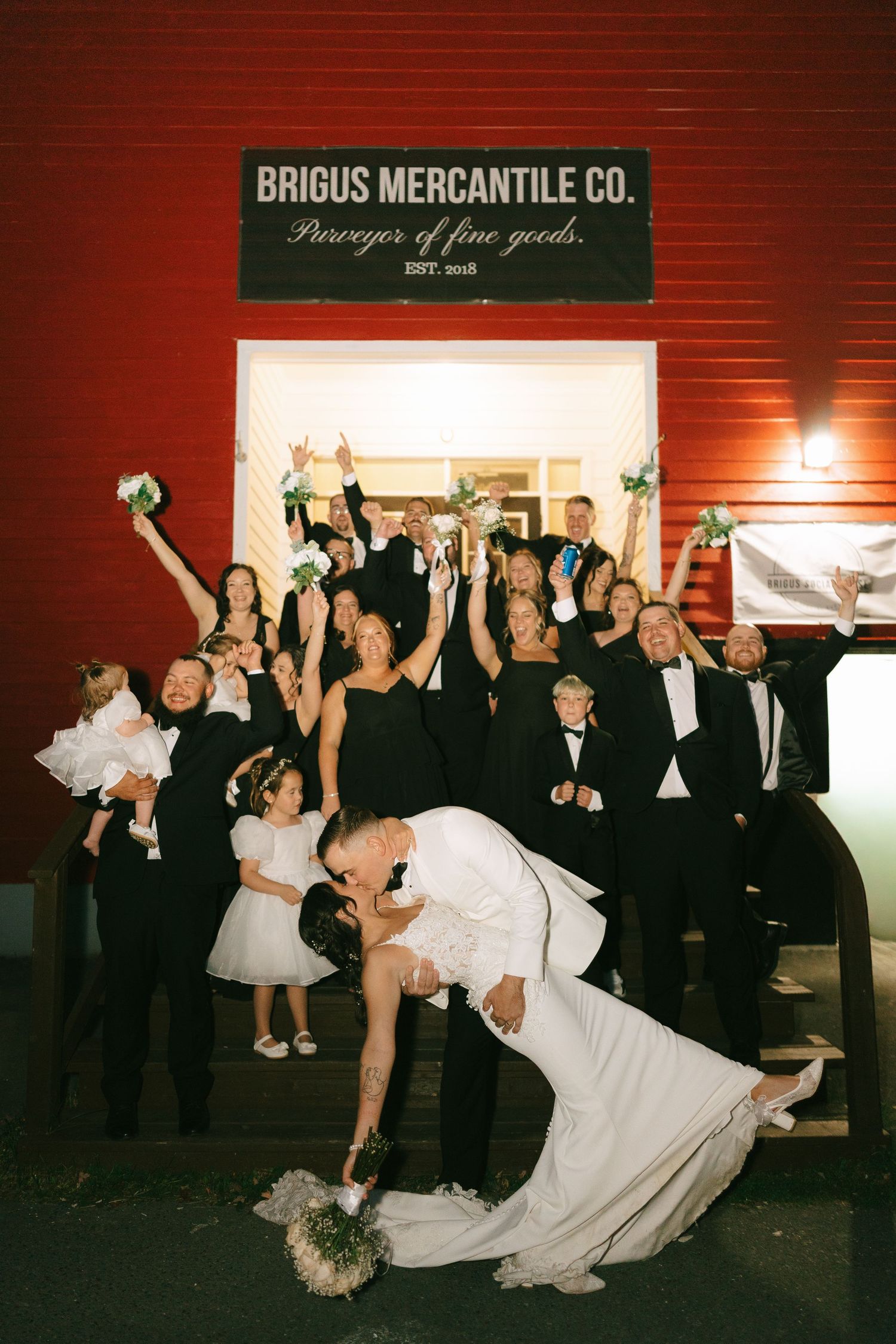 A wedding party celebrates with joyful poses and dance moves outside a rustic Briggs Mercantile Co. storefront at night.