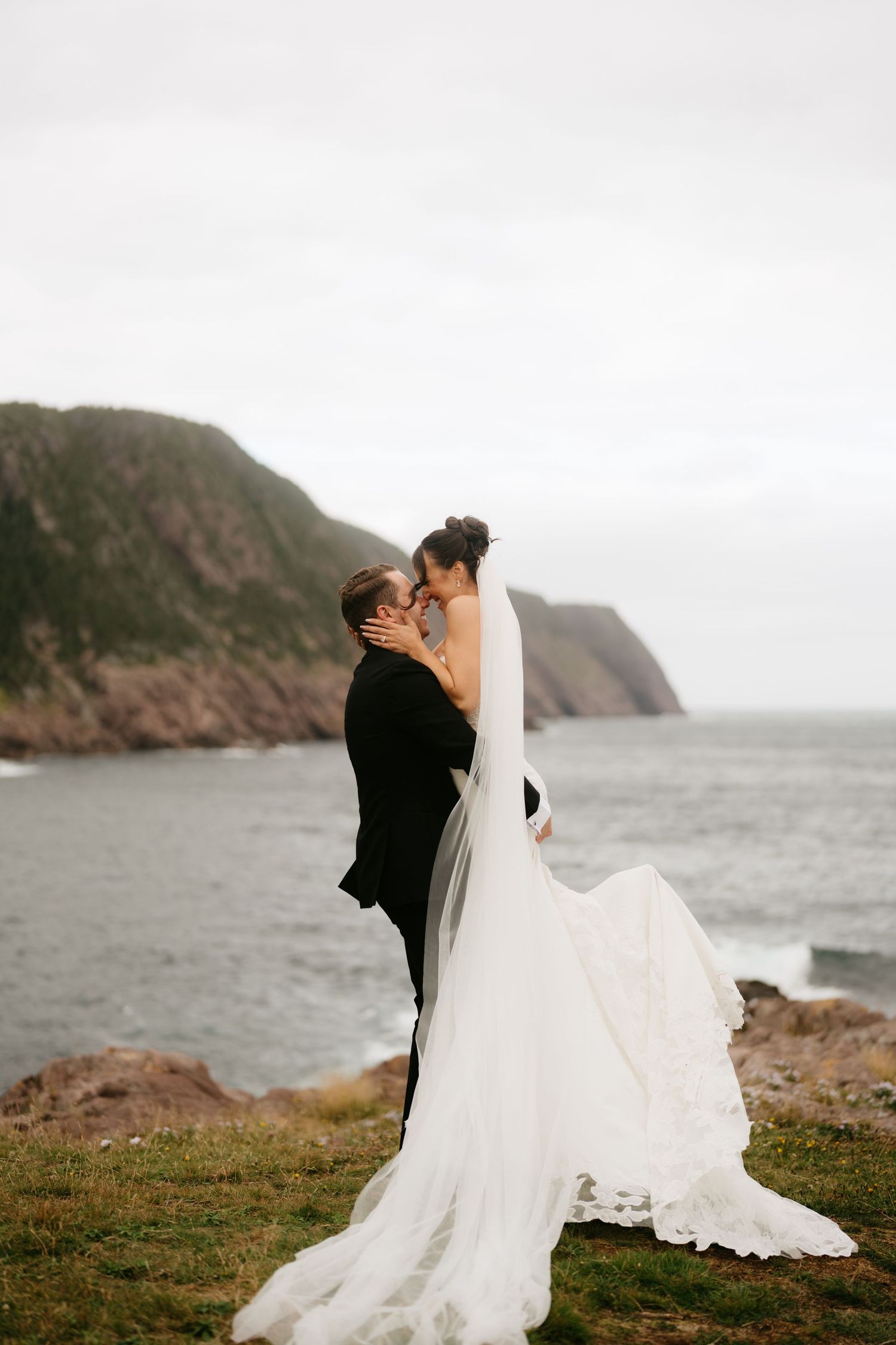 Bride and groom share a romantic kiss on a coastal cliff, with ocean waves and green hills in the background.