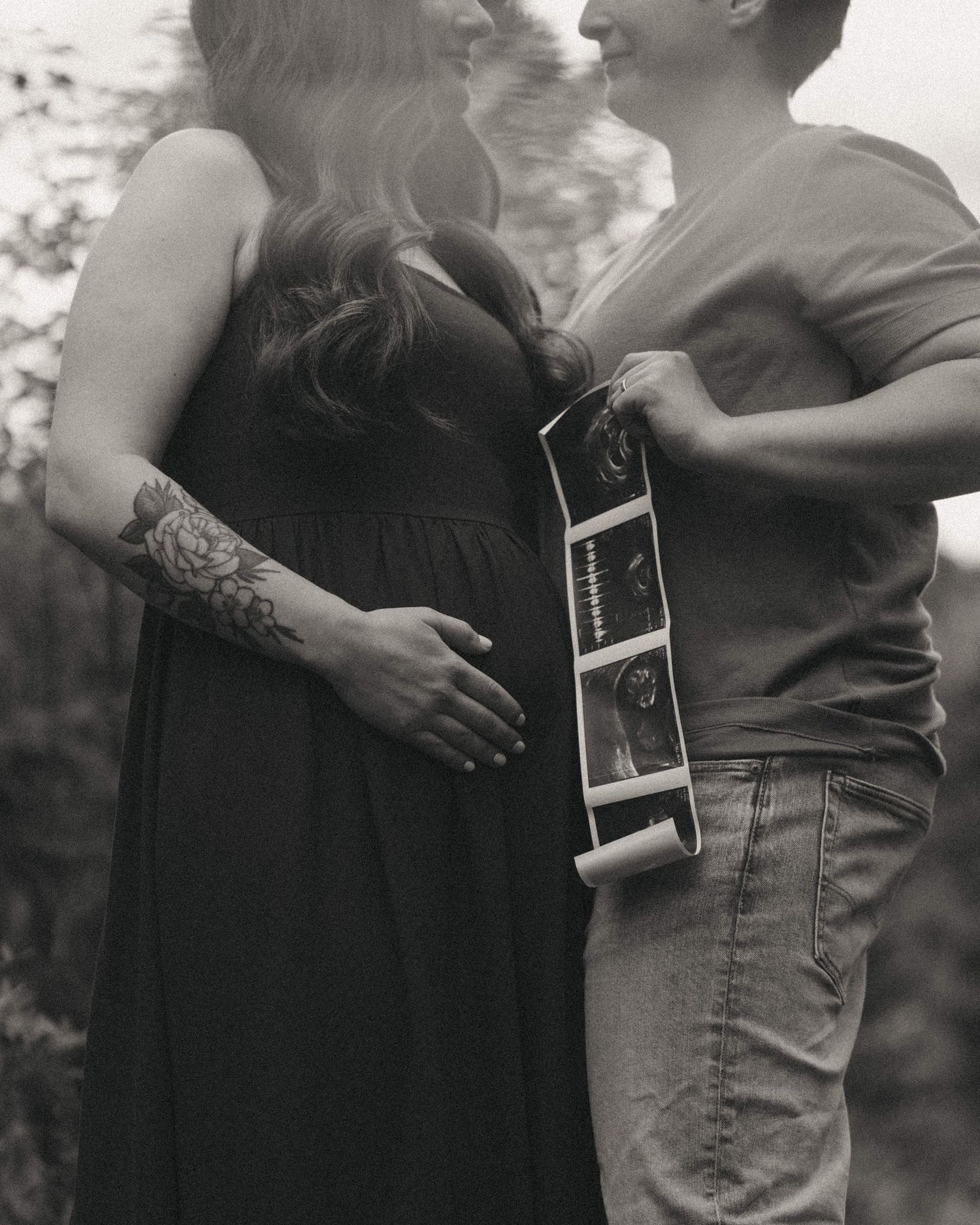 Black and white maternity photo of couple embracing outdoors, woman in black dress, man holding ultrasound photos.