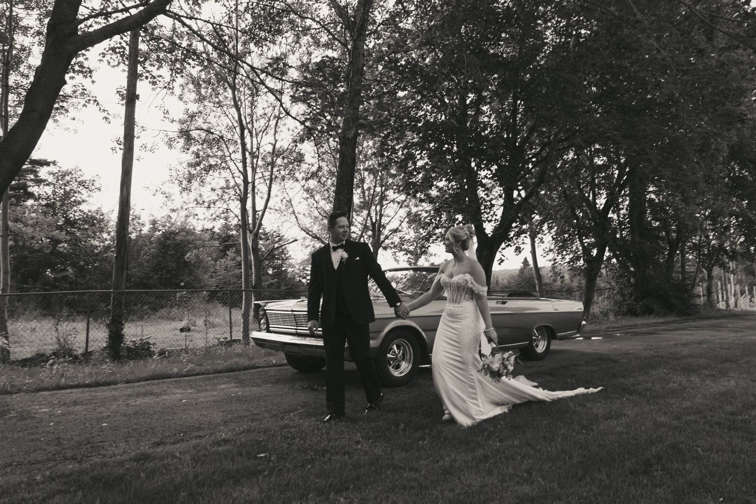 Black and white photo of a bride and groom posing by a vintage car on a grassy lawn surrounded by trees.