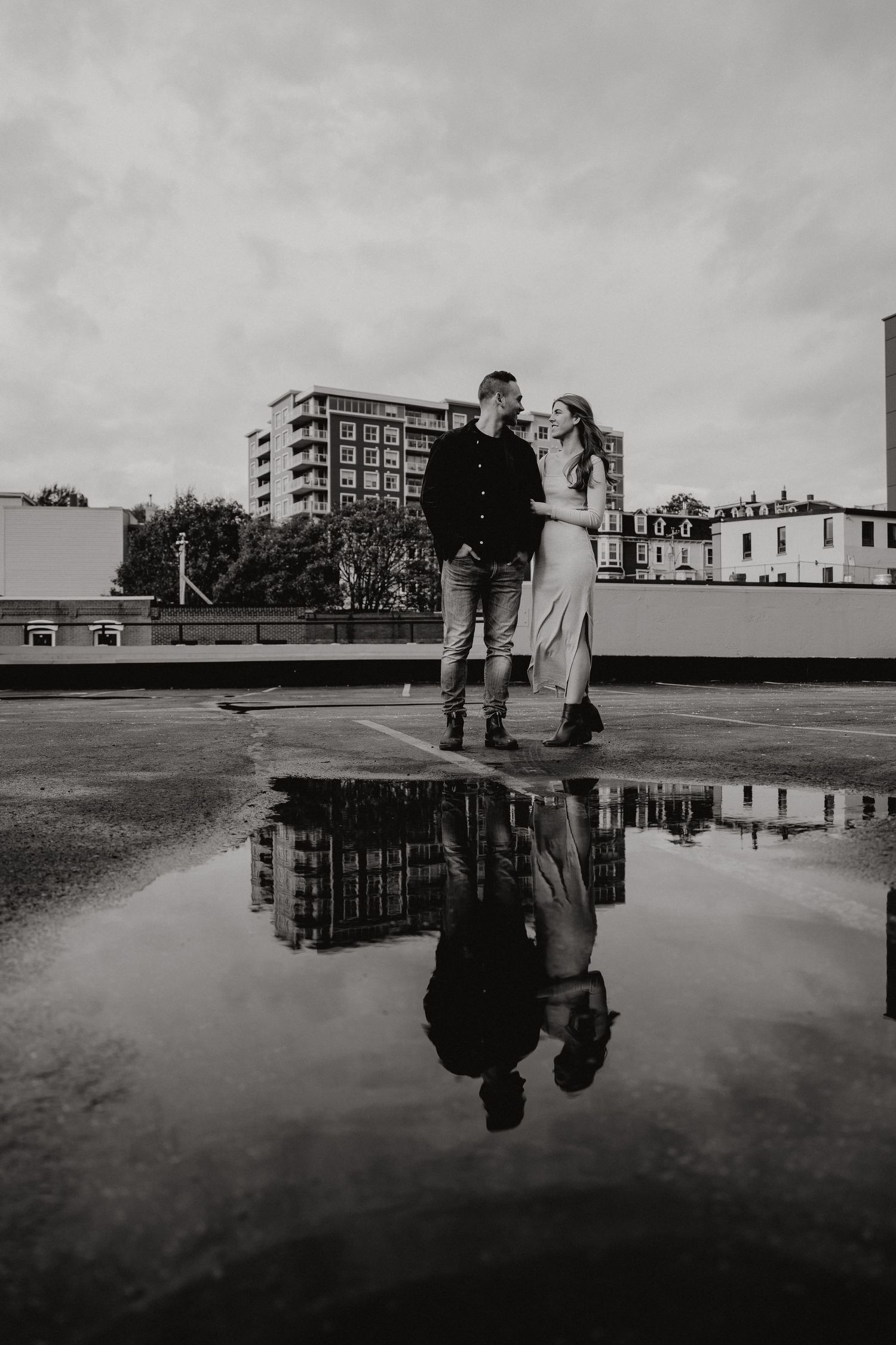 Black and white photo of two people reflected in a rooftop puddle with urban architecture behind them.