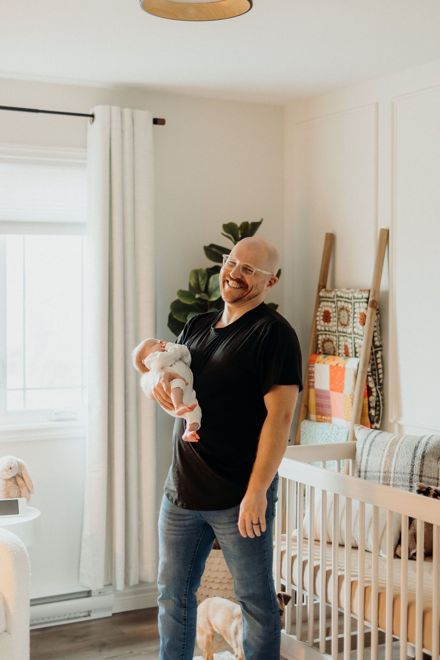 Adult wearing black t-shirt plays with baby near white crib in sunlit room.