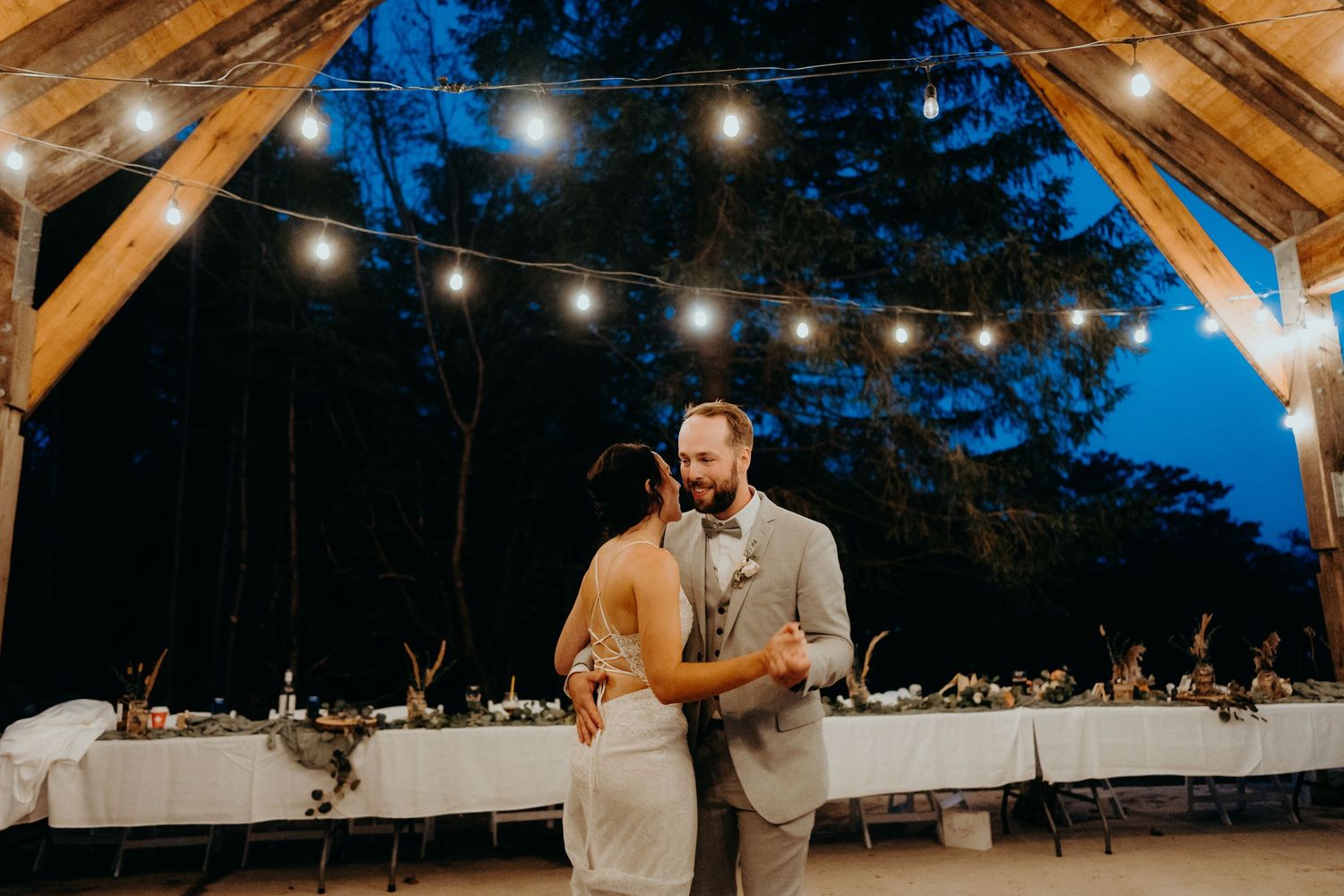 Reception dinner setup under wooden pavilion with string lights at dusk.
