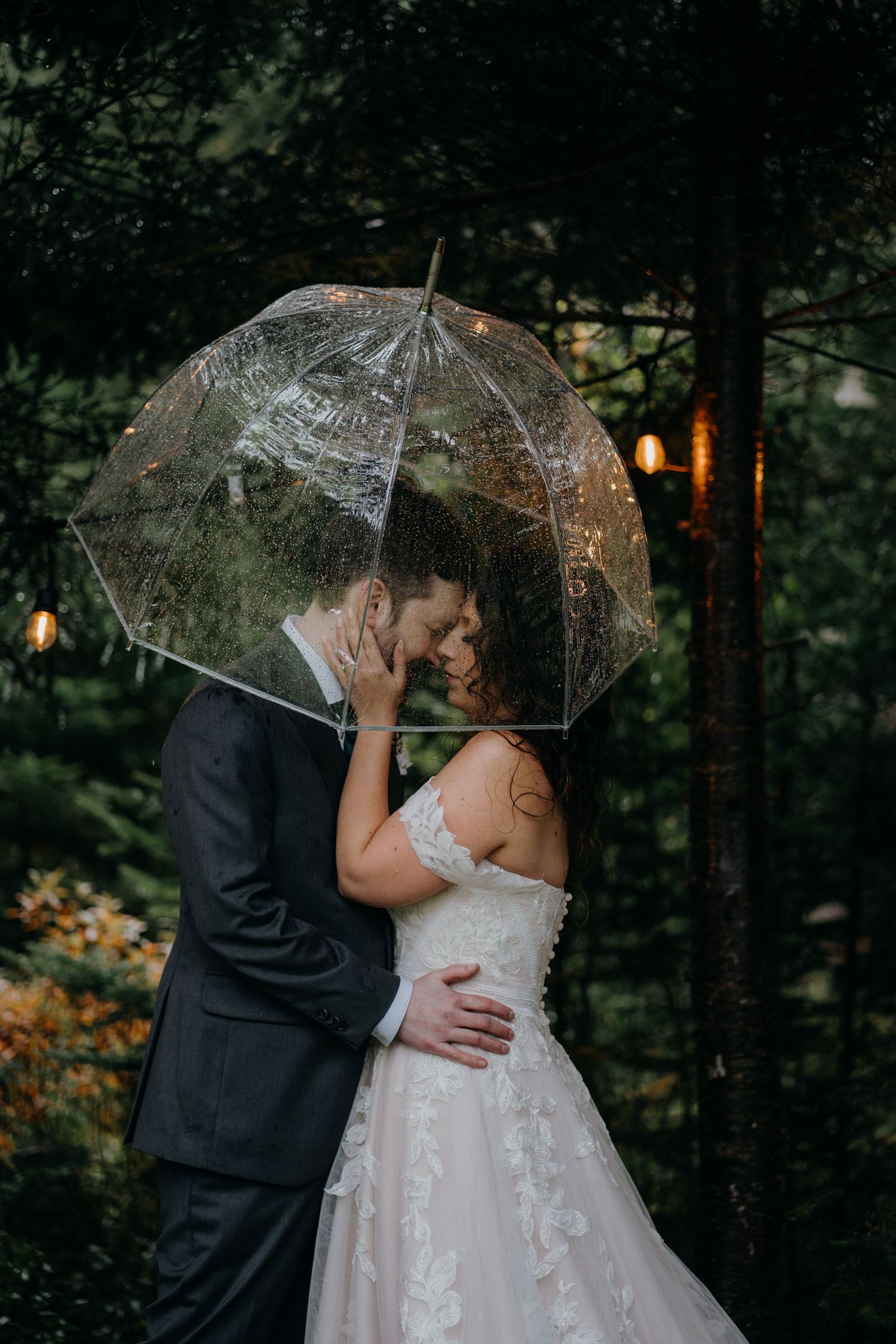 A romantic wedding couple shares an intimate moment under a clear umbrella in a moody forest setting.