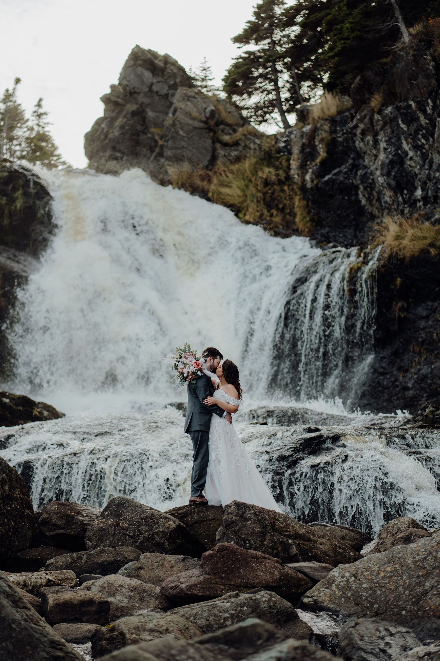 A couple embraces in front of a cascading waterfall surrounded by rocky terrain in a romantic outdoor setting.