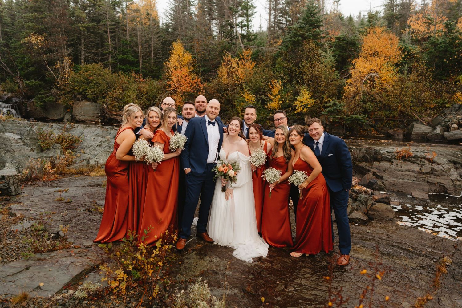 Wedding party poses together against autumn forest backdrop with vibrant orange foliage and rocky terrain.