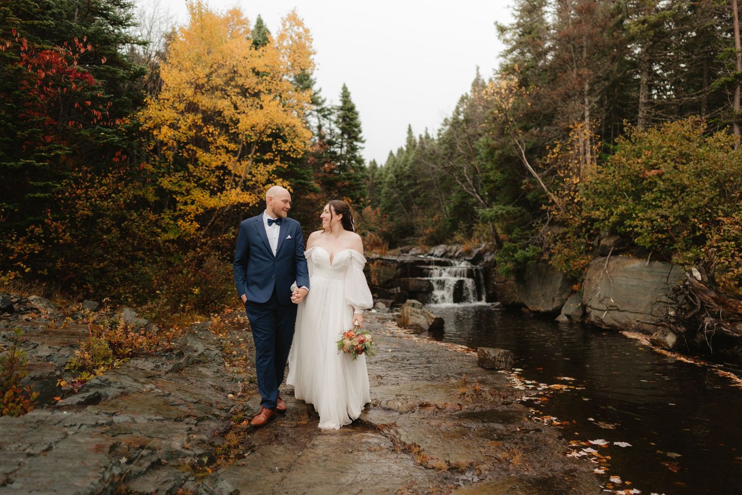 A couple in formal wedding attire stands together among autumn foliage and cascading waterfalls in a scenic forest setting.