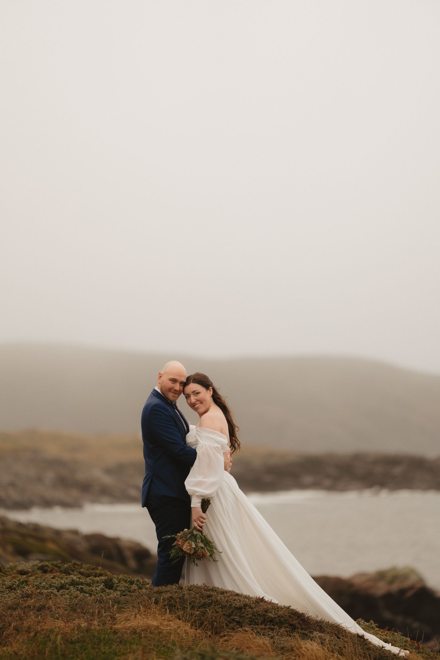 A romantic wedding couple embraces on a misty coastal cliff overlooking the ocean at sunset.
