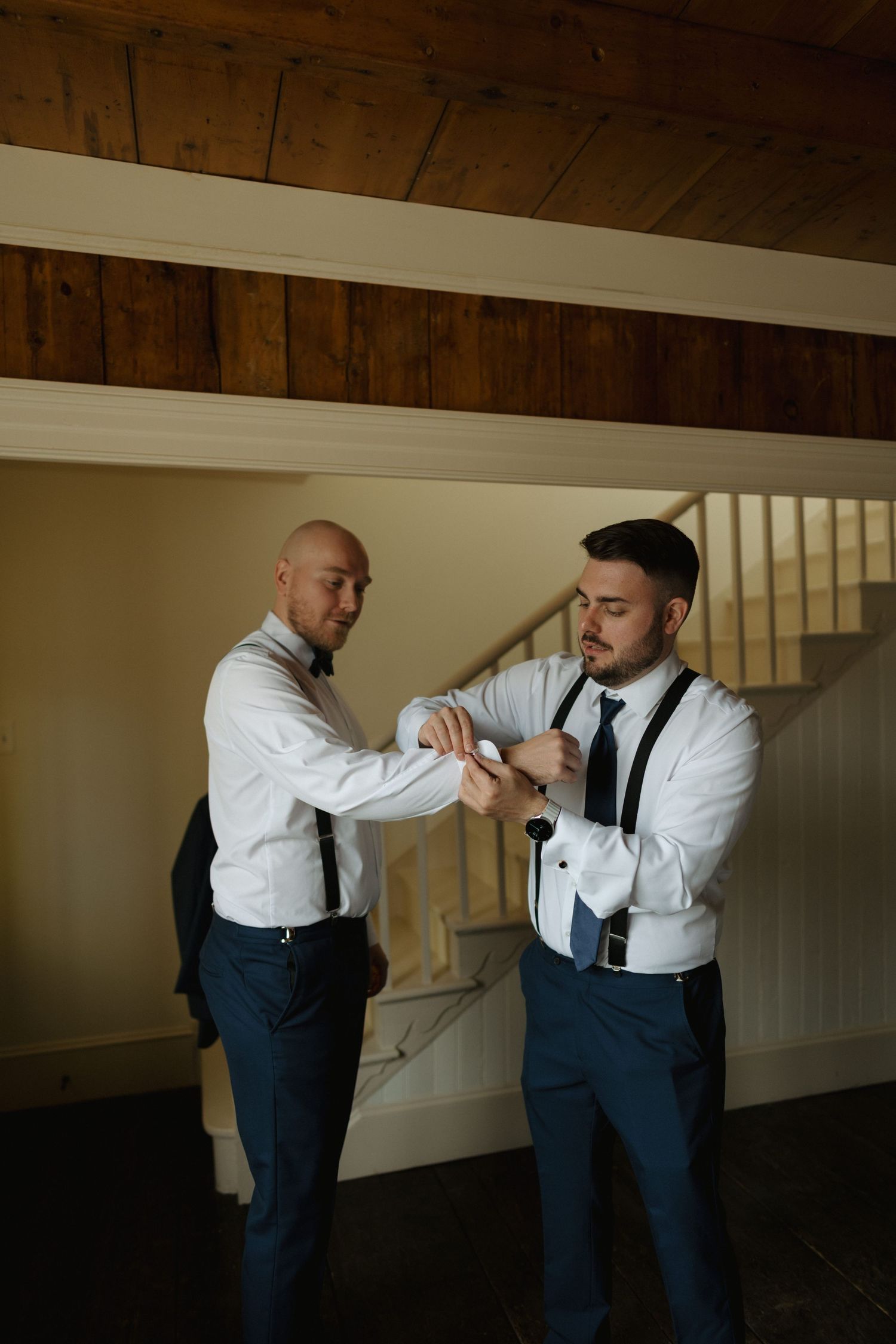 Two men in formal attire with suspenders and ties interact in a dimly lit interior space.