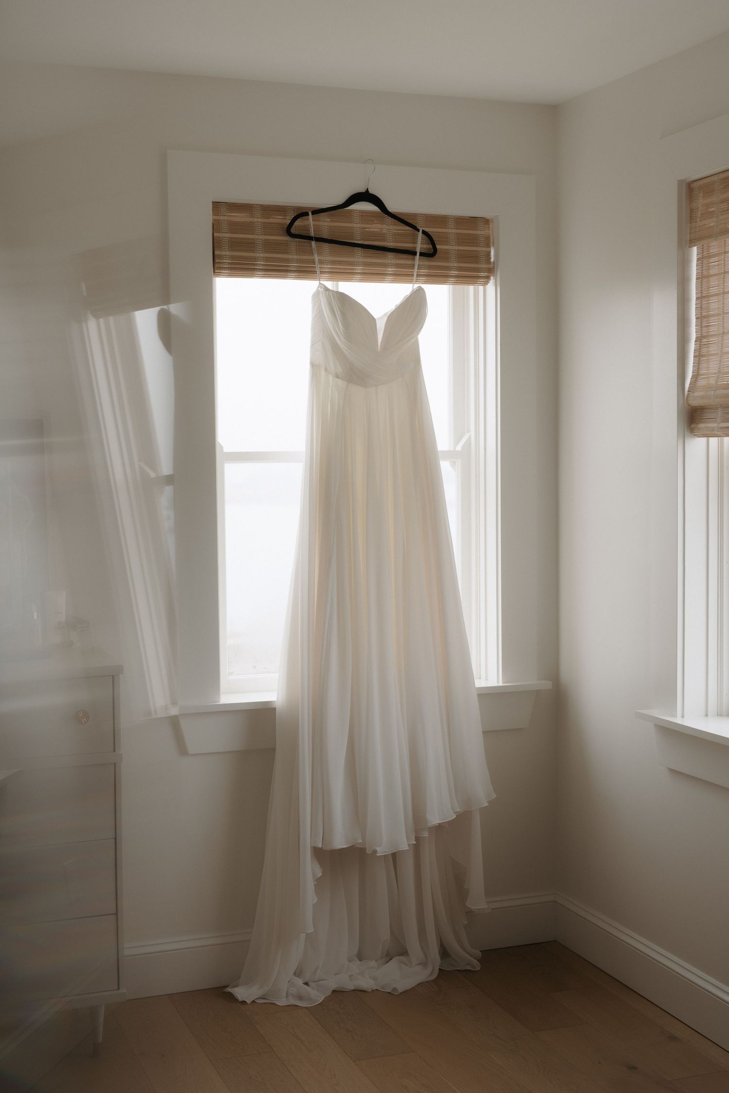 A series of white wedding dresses hanging in a bright sunlit room with bamboo window shades.