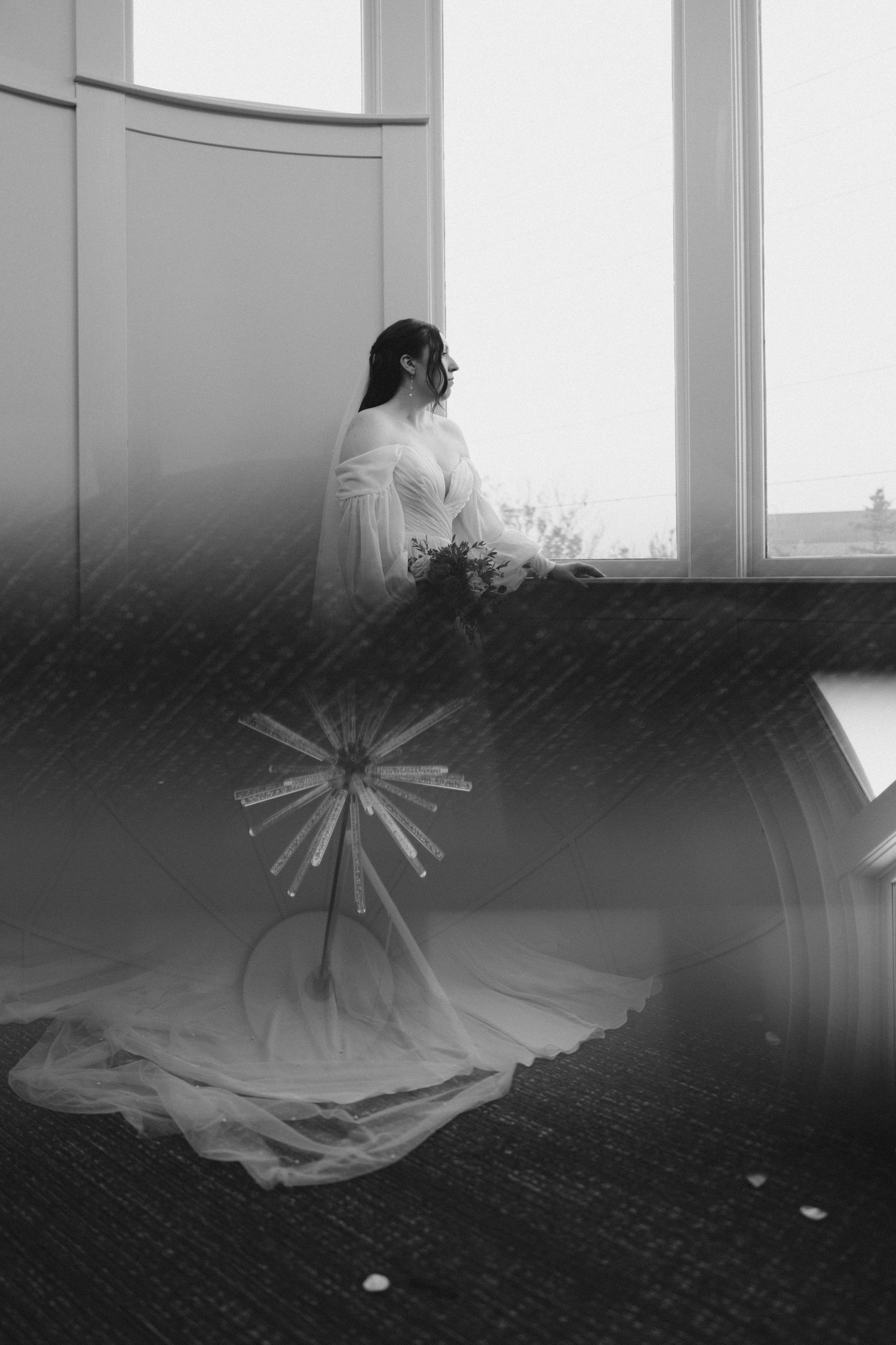 A black and white artistic portrait of a person sitting by a window with a starburst reflection on the floor.