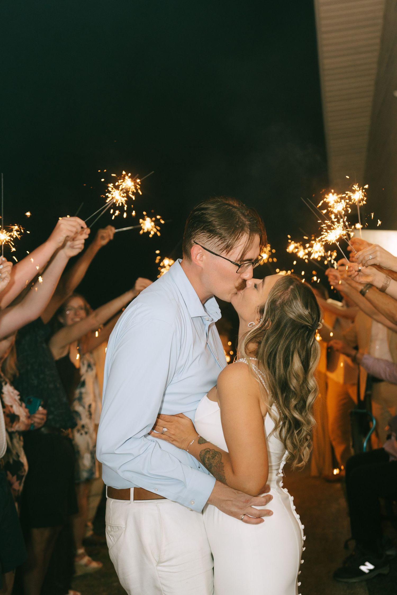 Newlywed couple sharing a kiss during a sparkler send-off at their outdoor evening wedding reception.