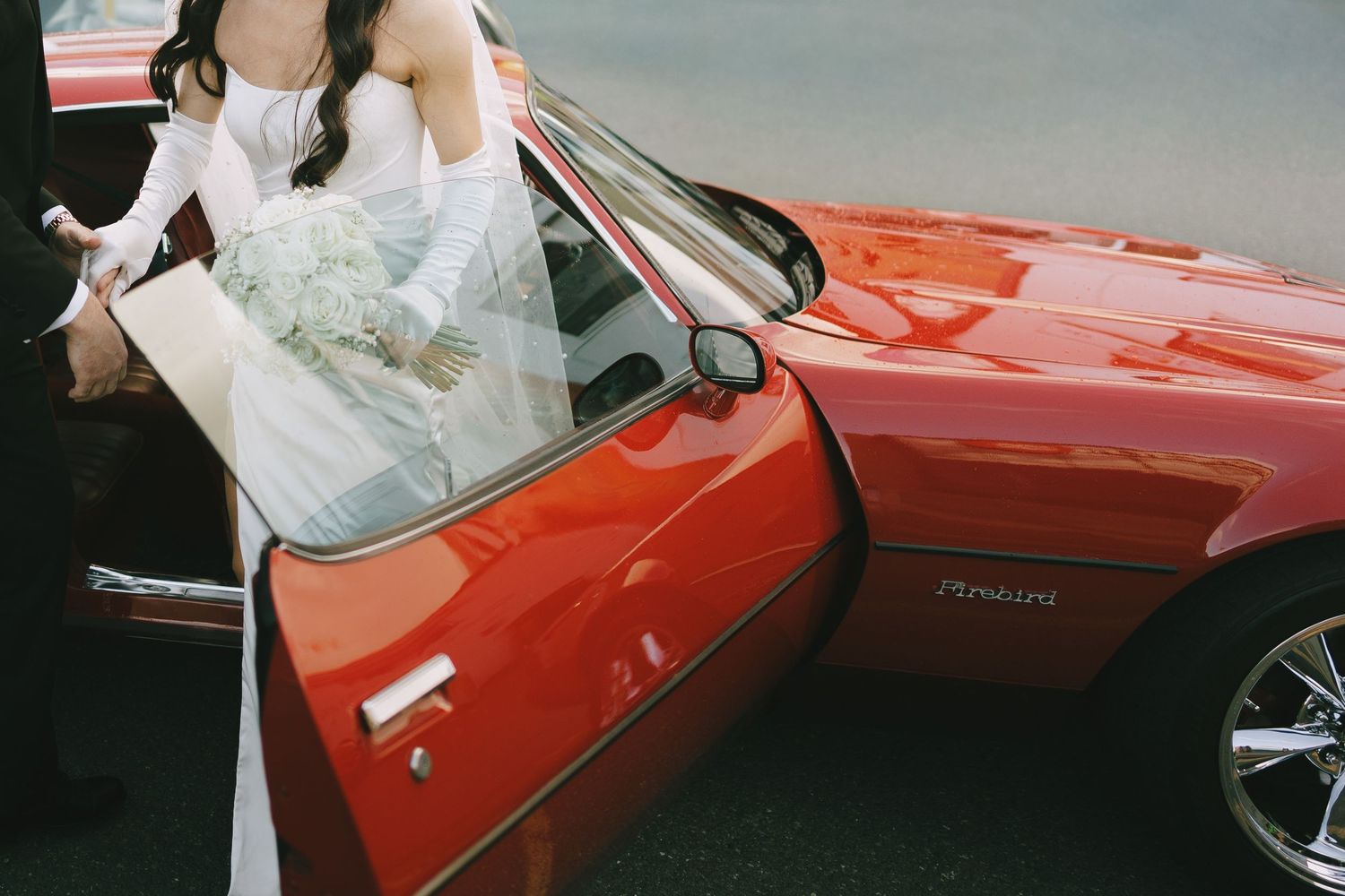 Bride in white wedding dress stepping out of a classic red convertible sports car on her wedding day.
