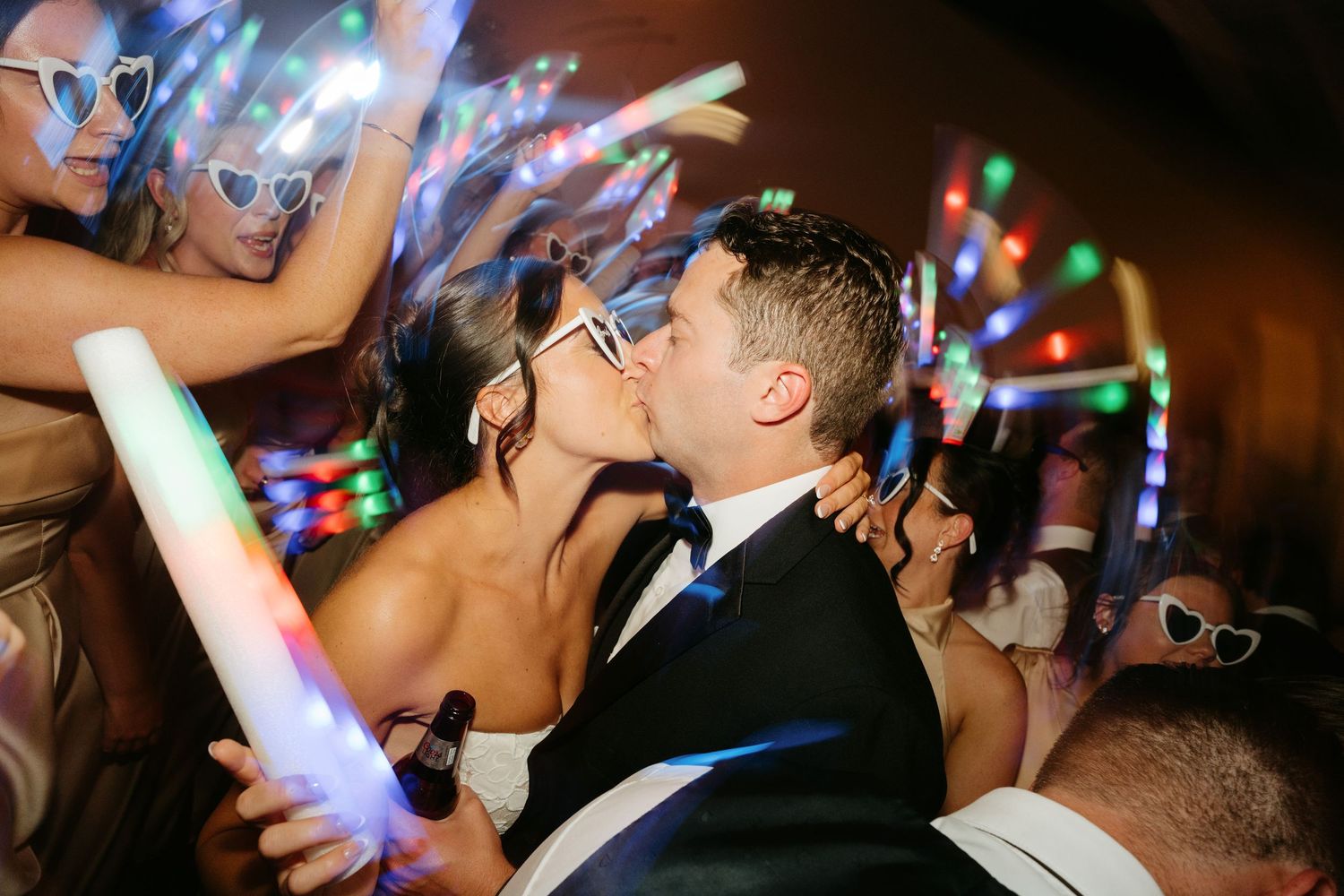 Newlyweds share a kiss on the dance floor surrounded by guests waving colorful LED foam sticks at a wedding reception.