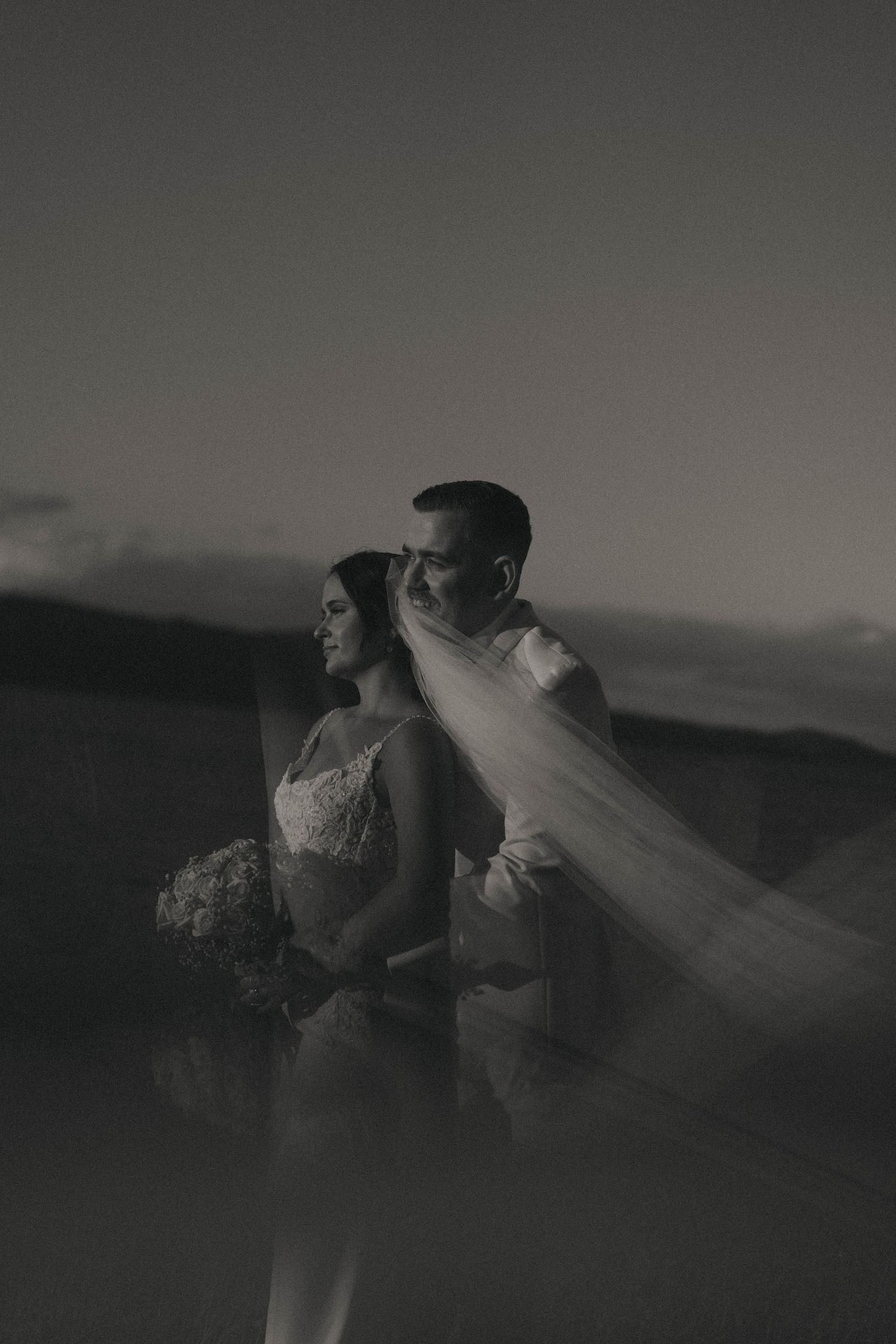 A romantic black and white wedding photo series showing a couple in formal attire with a flowing veil against a dramatic sky.