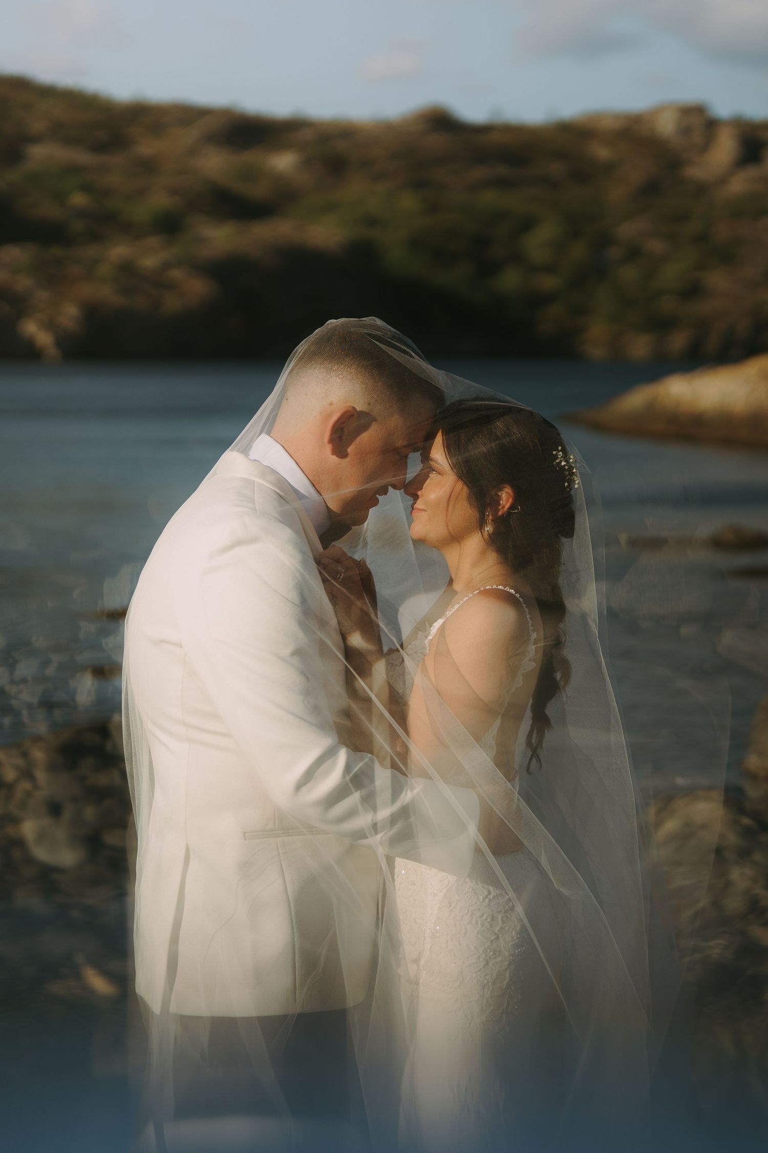 Romantic couple in white attire share intimate moments by a lakeside during sunset photoshoot.