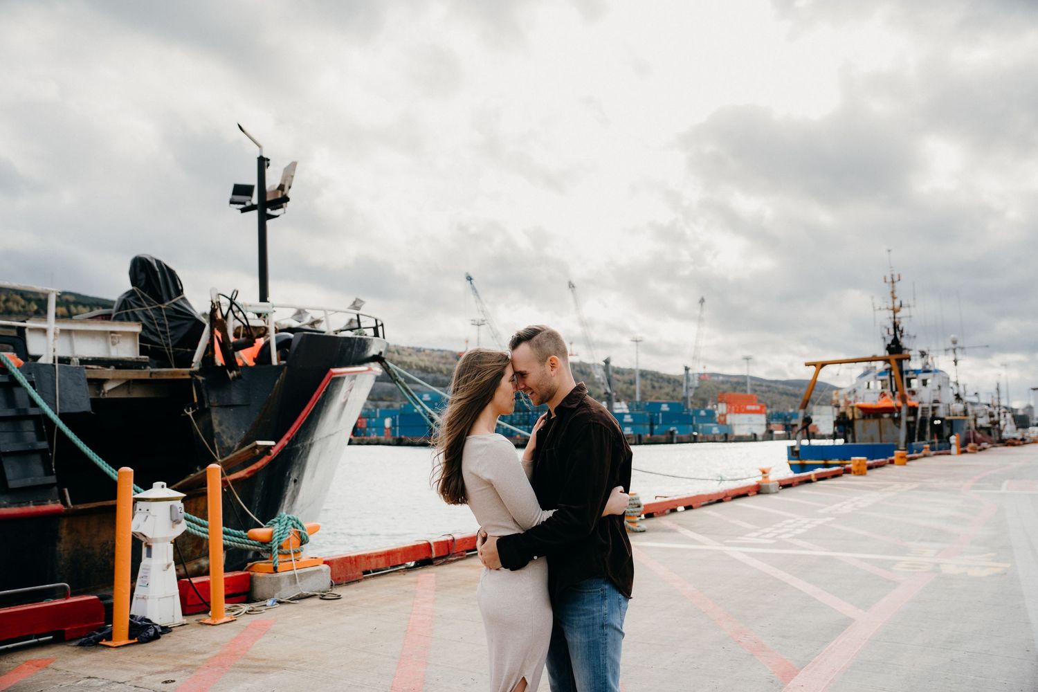 A couple embraces while standing on a waterfront pier with boats in the background.
