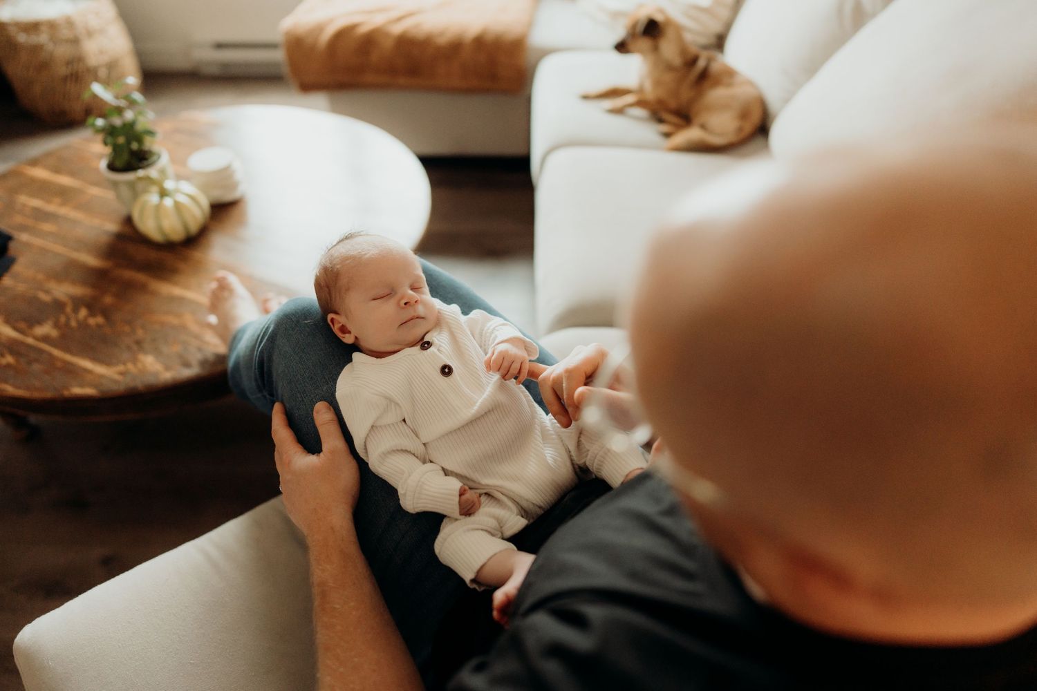Gentle hands holding a newborn wearing a white ribbed outfit in a warm indoor setting.