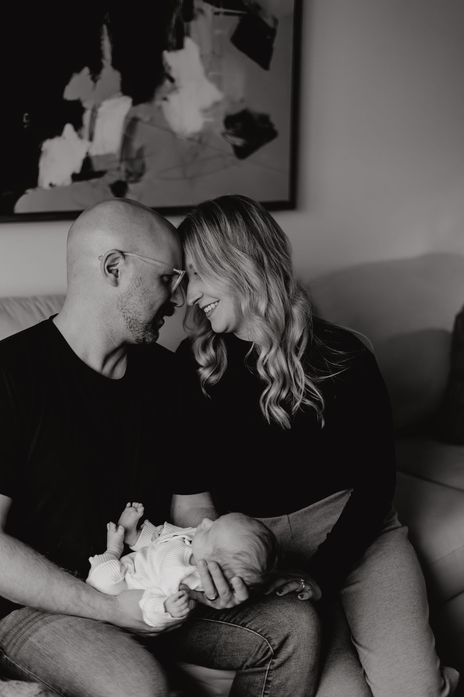 Moody black and white photograph of parents sharing tender moment with baby on couch.