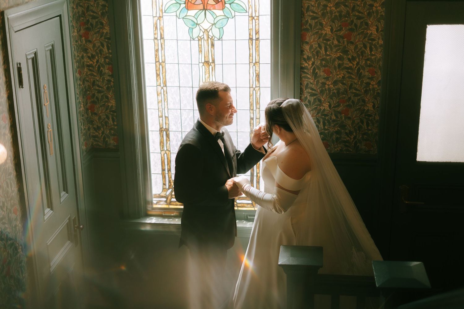 Couple standing in church interior with stained glass windows and vintage wallpaper backdrop.