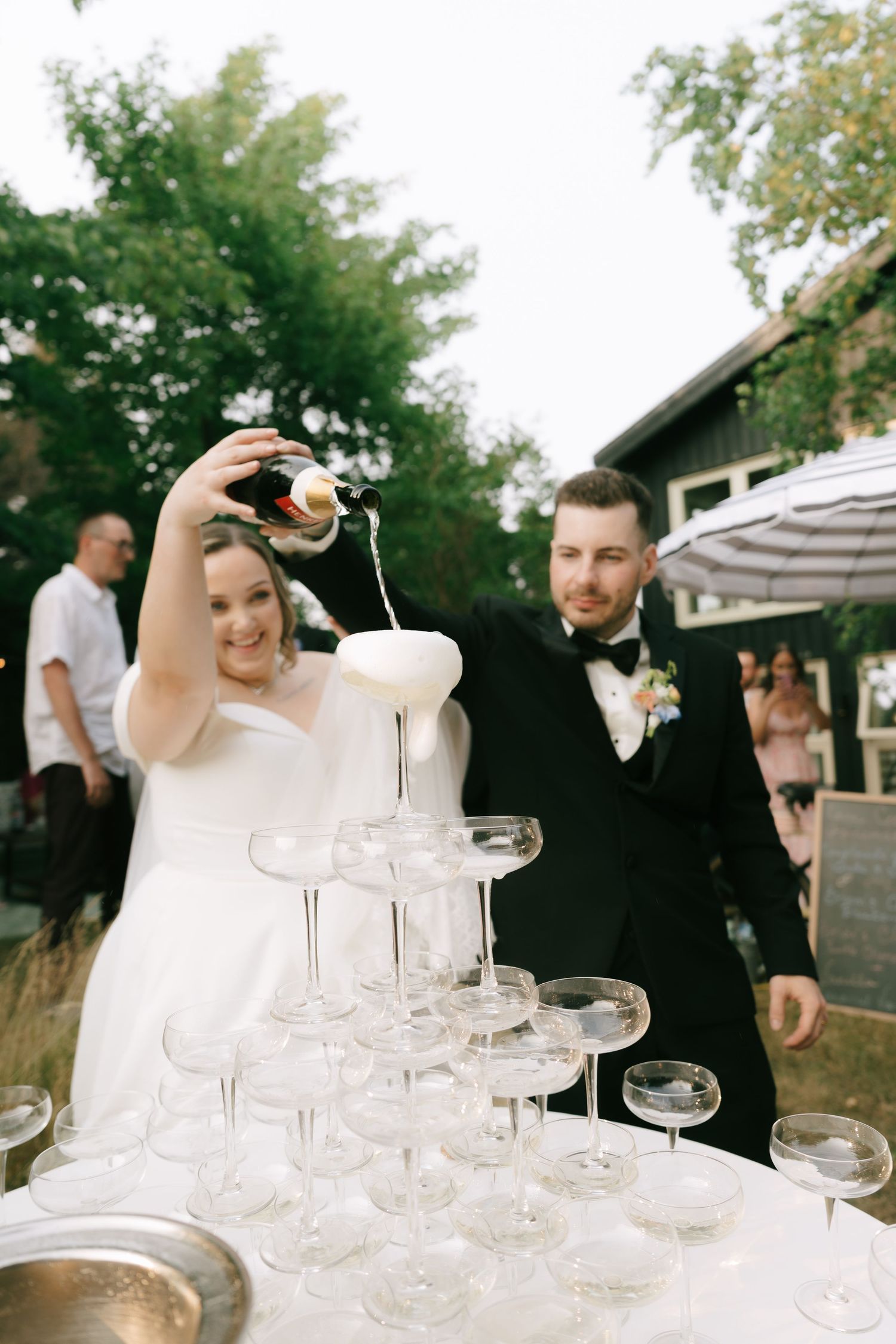 Guests pouring champagne into a tower of crystal glasses at an outdoor reception.