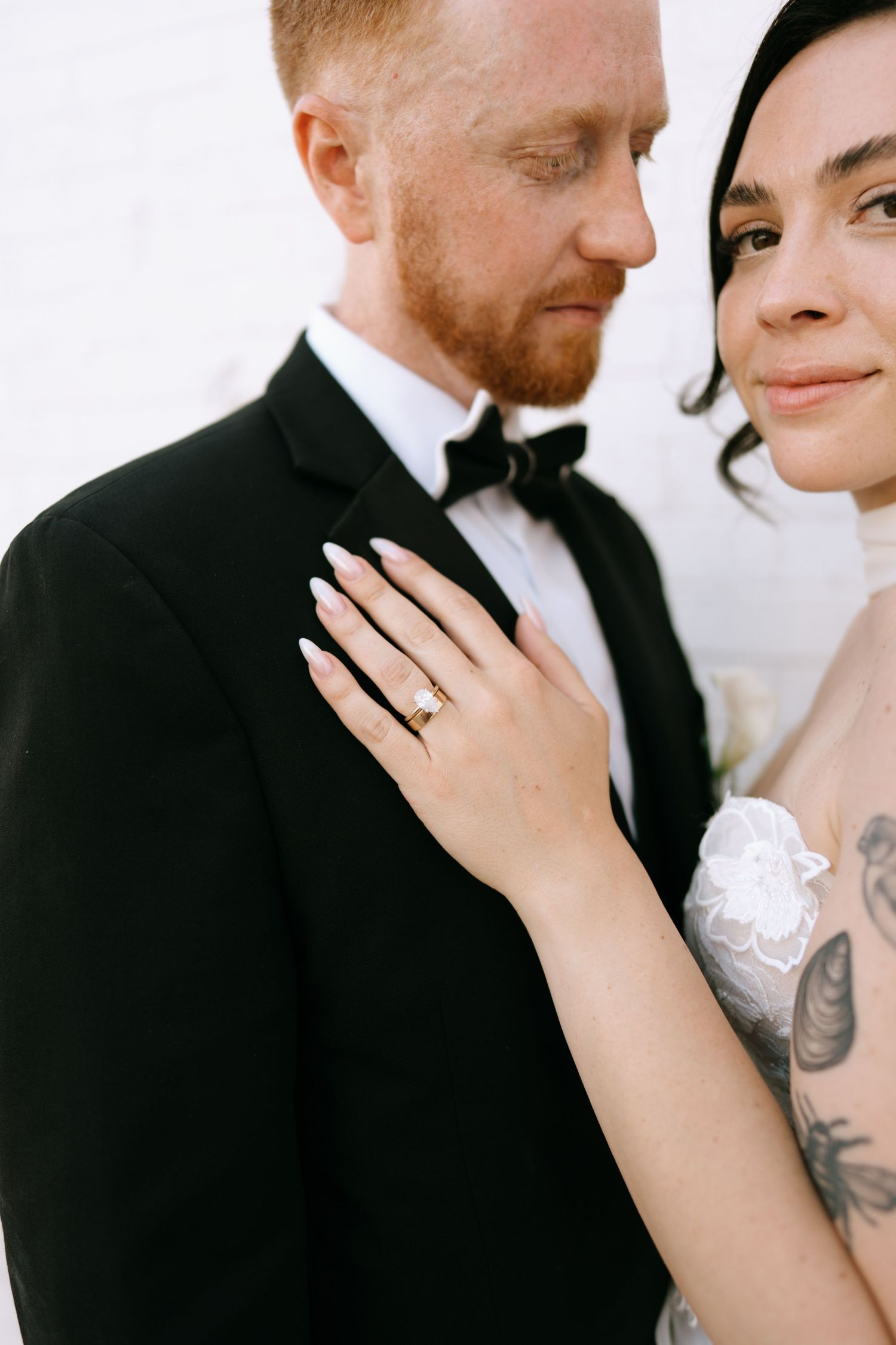 Close-up of bride's hand with diamond engagement ring resting on groom's black tuxedo lapel.