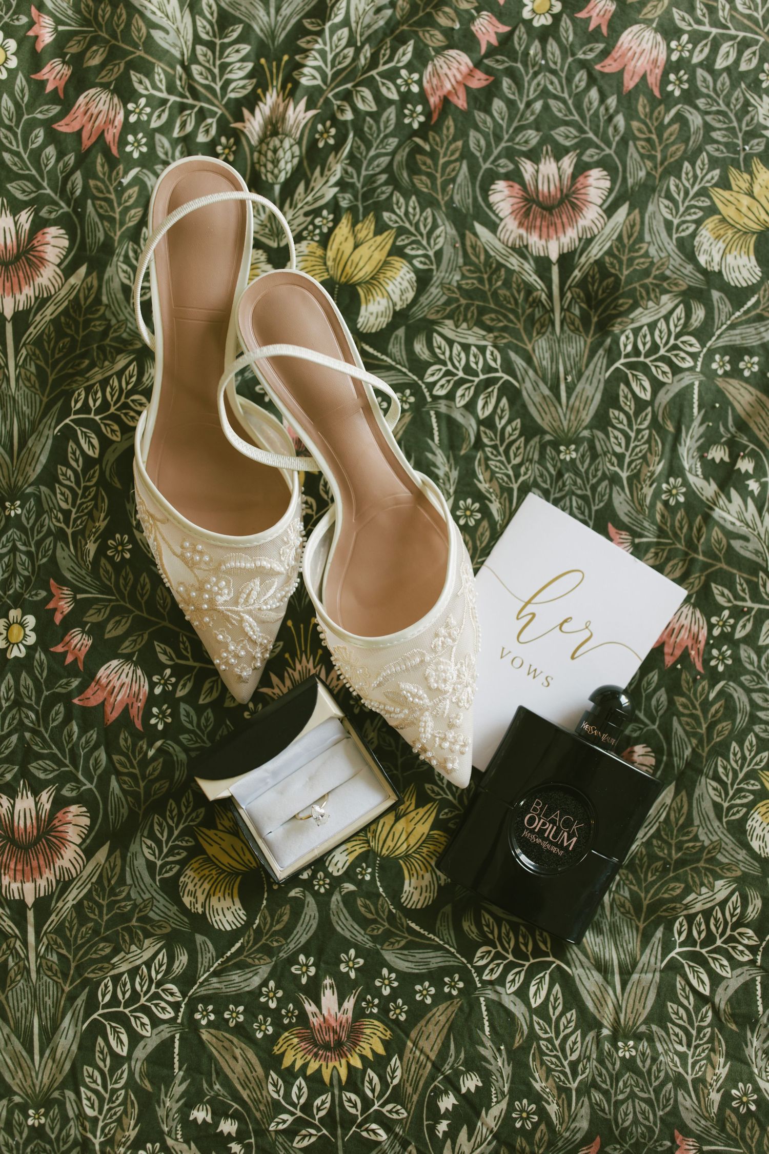 Bridal flat shoes with floral embroidery beside vow book, diamond ring, and Black Opium perfume on green botanical wallpaper.