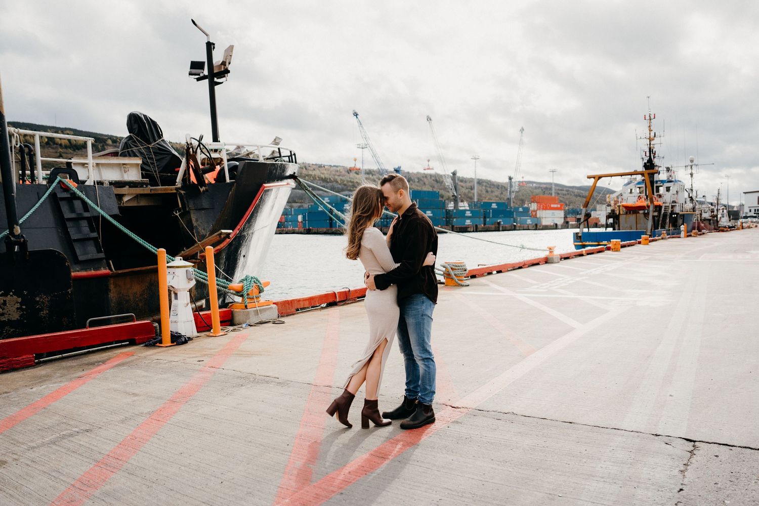 Couple in romantic pose on dock with fishing vessels and industrial equipment in background.