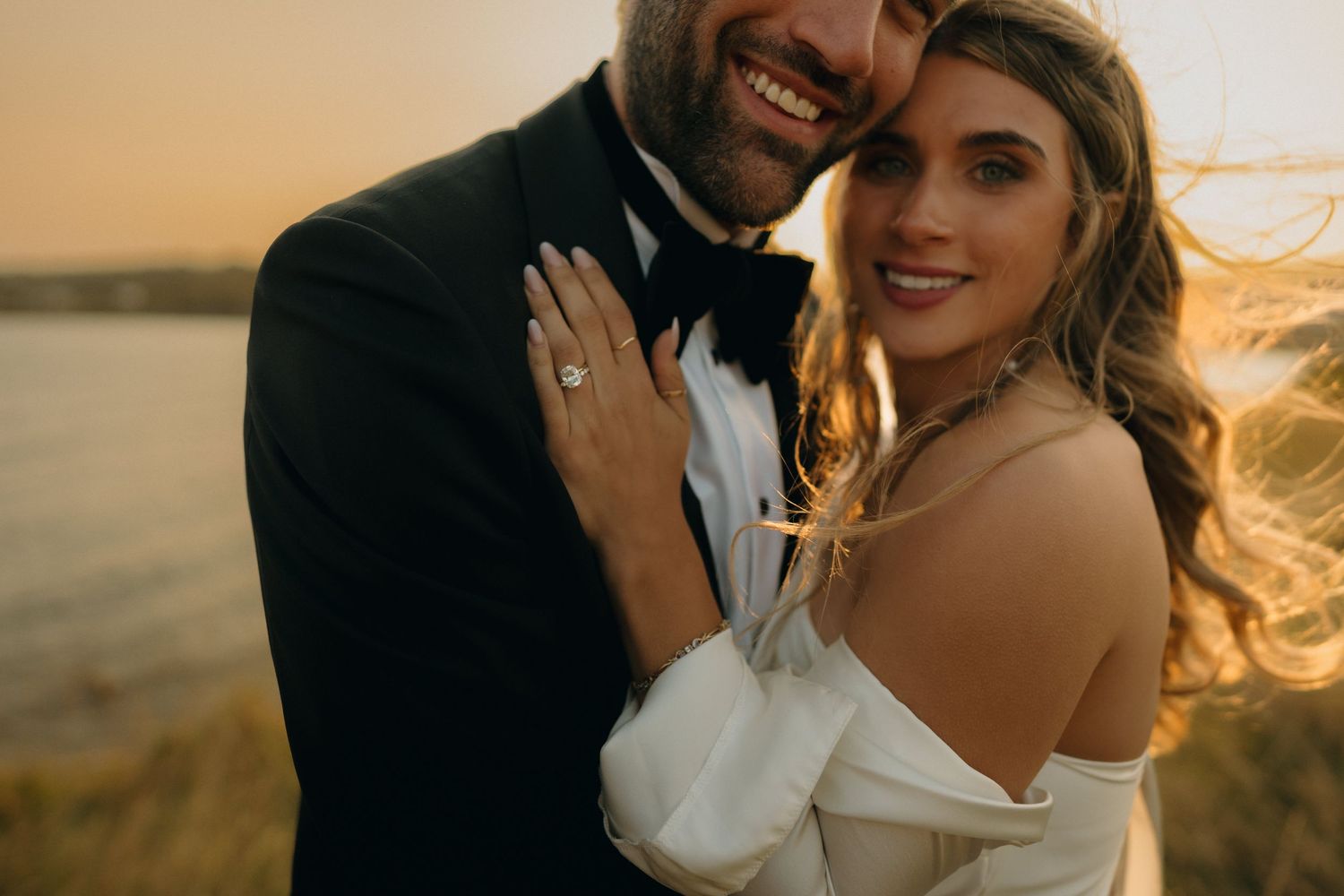 A romantic couple embraces during golden hour at sunset, with wind blowing through their hair.