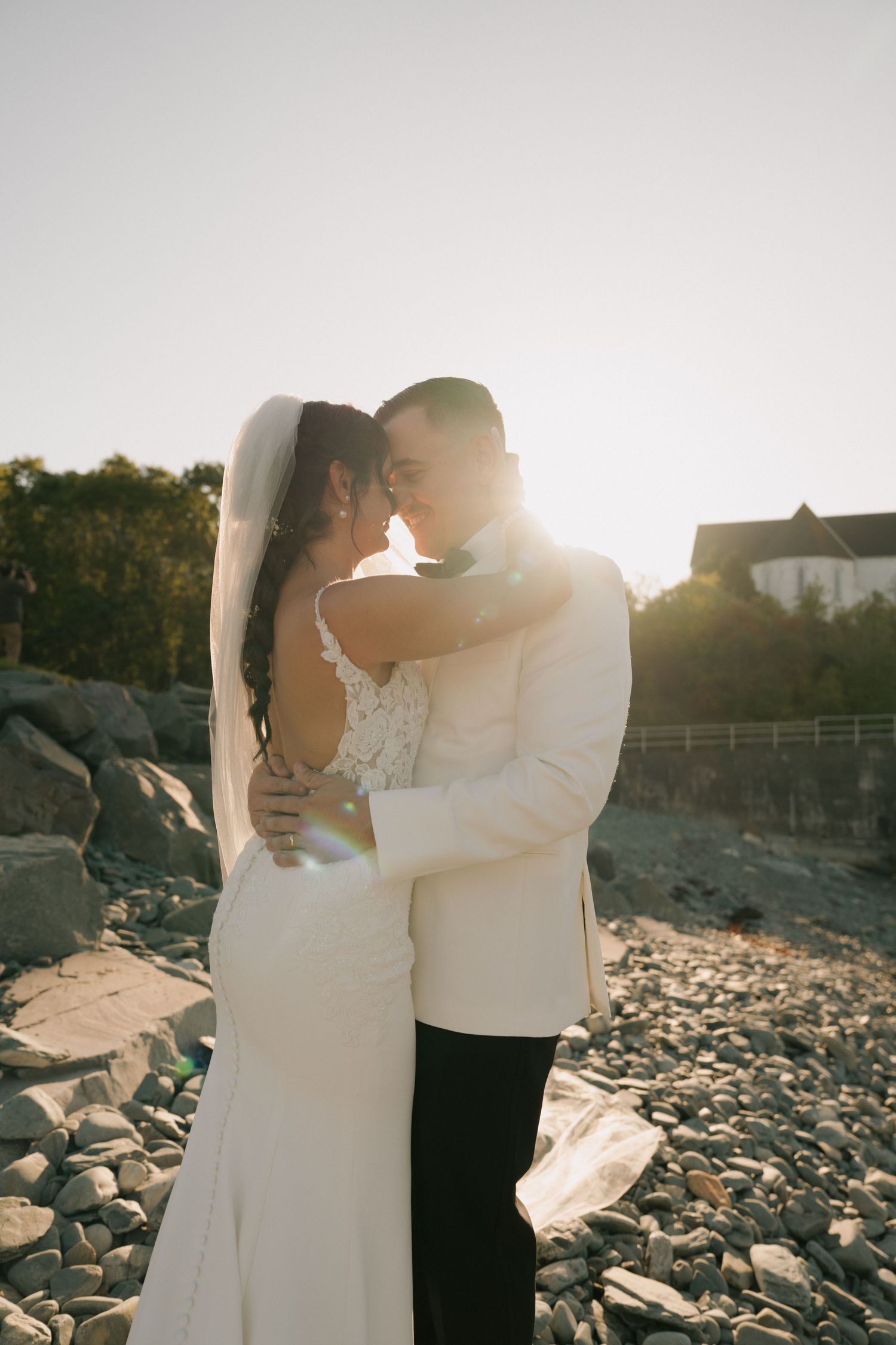 Romantic couple in wedding attire embracing on rocky beach during golden hour sunset.