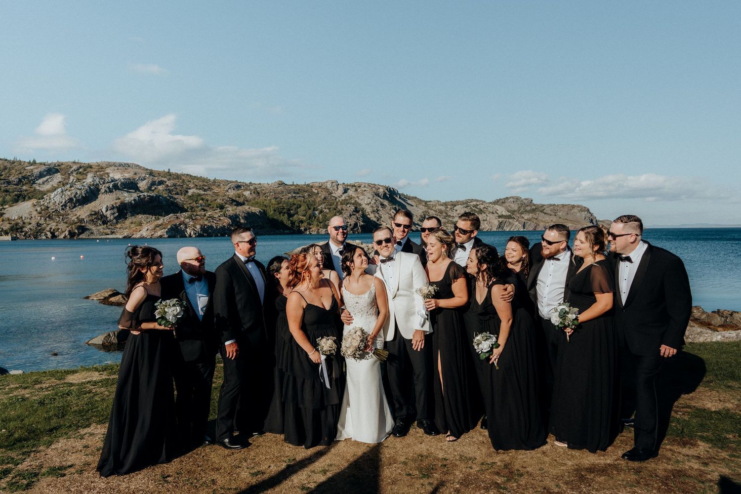 Wedding party stands together on scenic coastal cliffs overlooking blue ocean waters on a sunny day.
