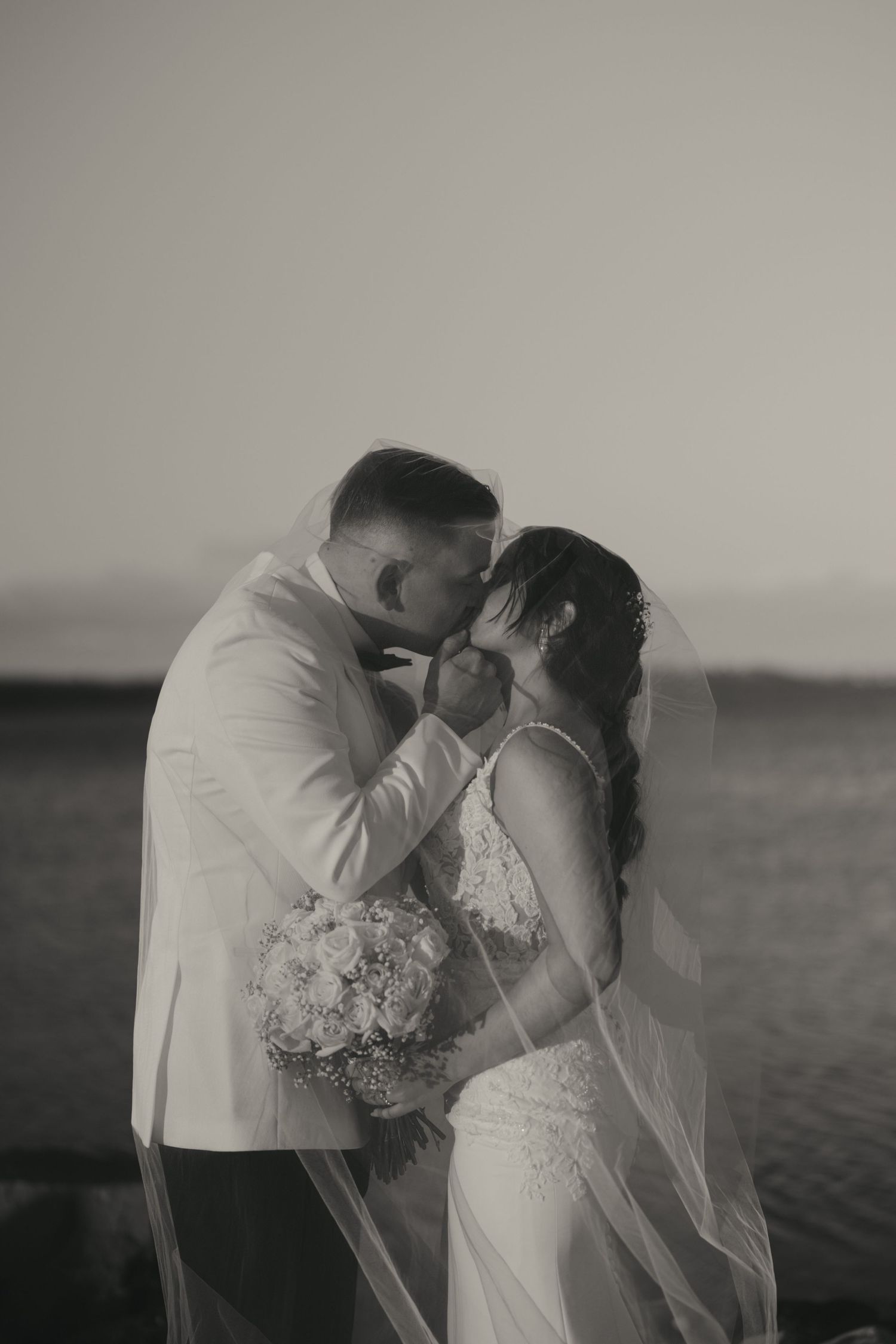 A romantic black and white wedding photo of a couple sharing an intimate moment by the water at sunset.