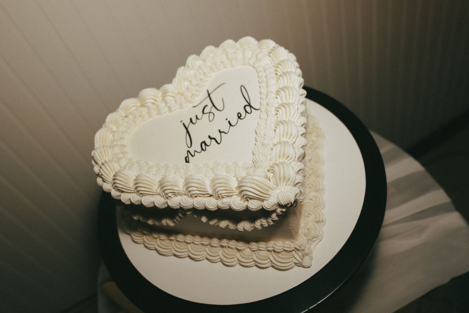 A white heart-shaped cake with delicate piped frosting decorations sits on a plate against a dark background.