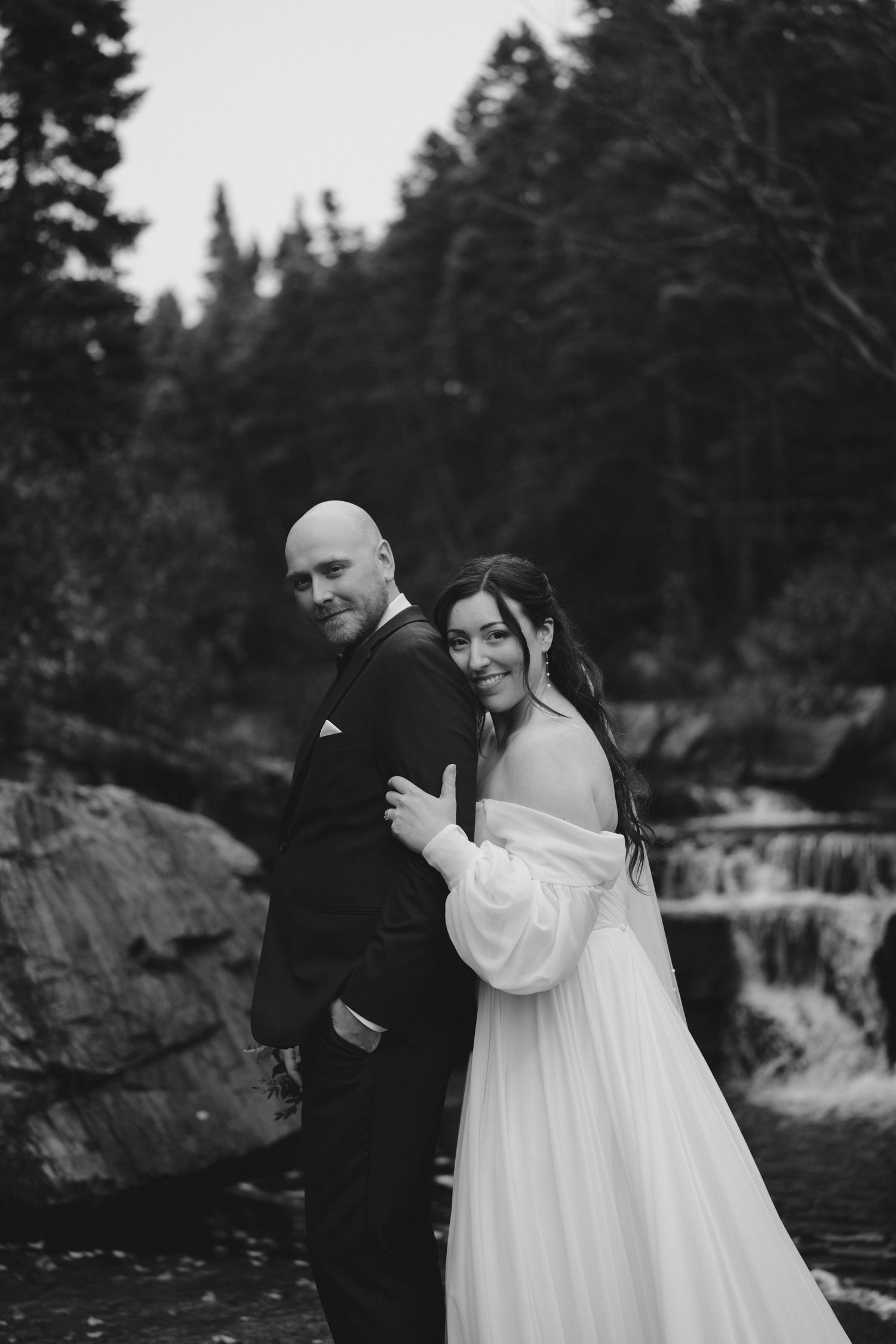 Black and white wedding portrait of a couple standing by a waterfall with evergreen trees in the background.