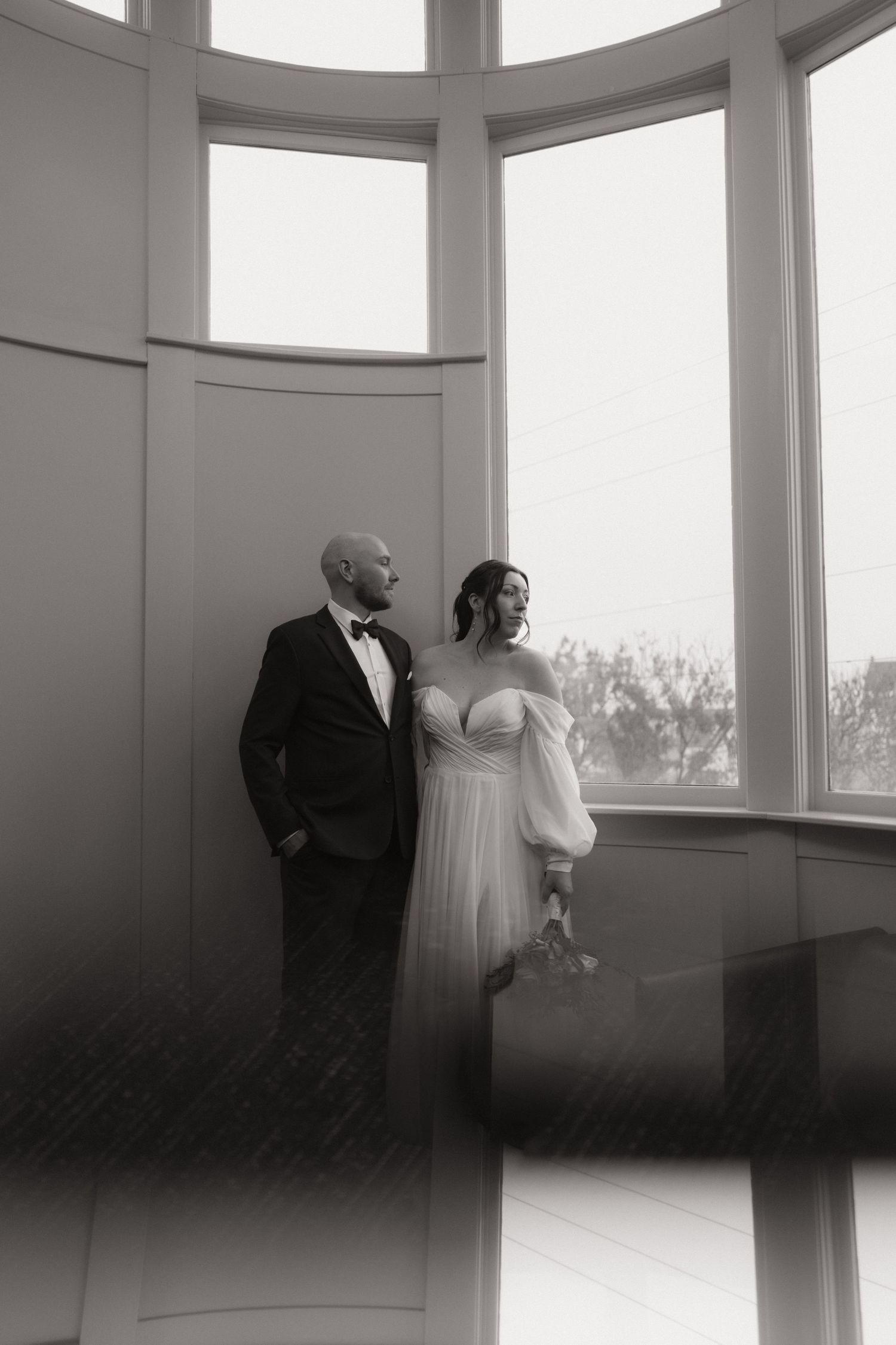 Black and white wedding photos show a couple in elegant attire standing by large windows in a bright room.