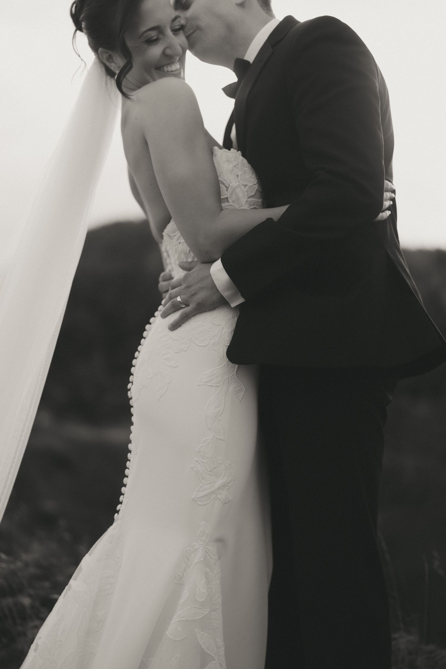 Black and white photo of a bride and groom embracing outdoors, her lace mermaid gown and veil flowing gracefully.